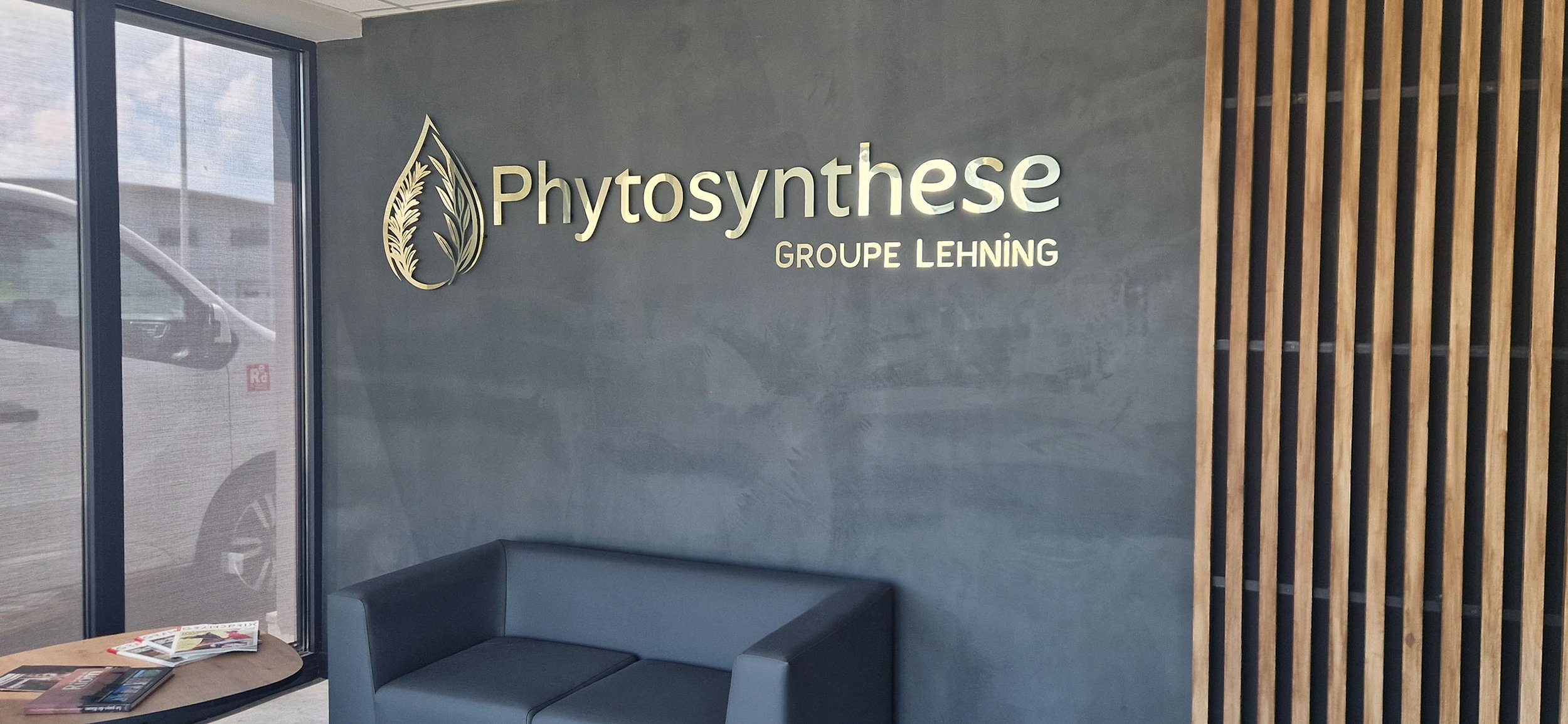 Une salle d'attente moderne avec un mur en béton gris, un logo en métal avec une feuille et des plantes, et un canapé gris foncé près d'une fenêtre.