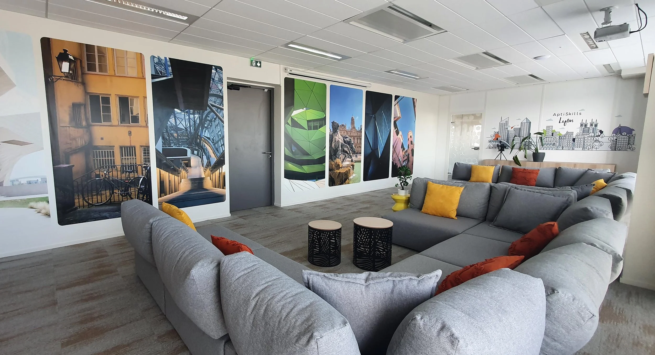 Salon avec un canapé gris, coussins orange et jaune, murs décorés avec des affiches de divers endroits, plantes en pot, et un mur avec une illustration de skyline.