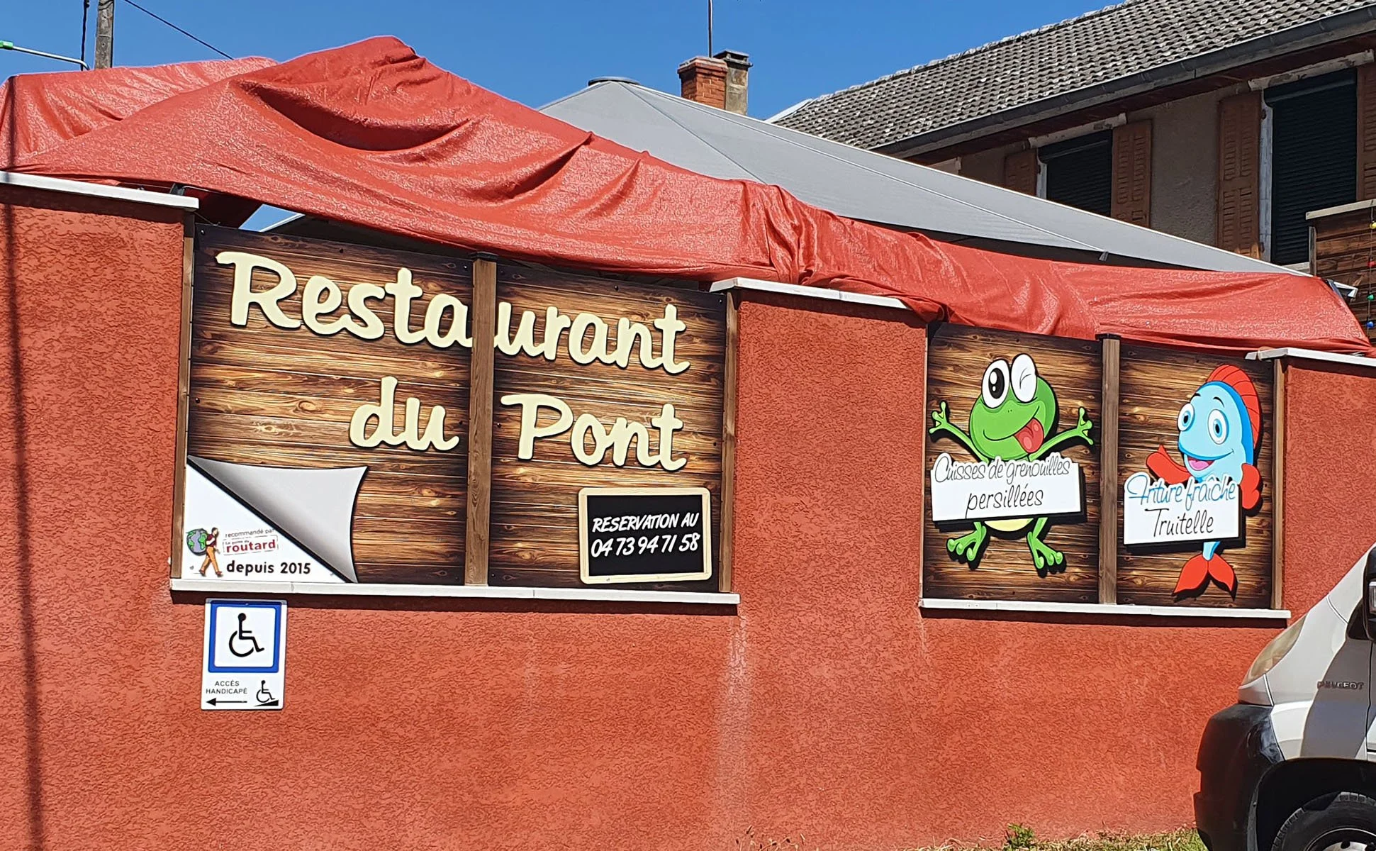 Une façade de restaurant du Pont avec des panneaux indiquant les spécialités de palourdes, crabs, et truite, décorés avec des dessins de crapaud et de poisson.