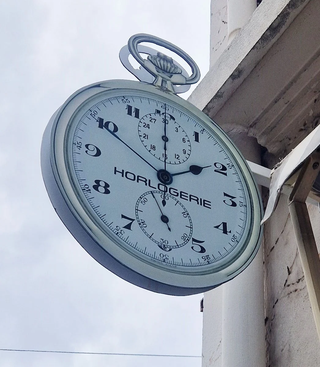 Horloge murale ronde de style vintage, affichant l'heure et mesurant le temps, fixée à un bâtiment, avec le nom 'HORLOGERIE' écrit au centre, contre un ciel gris.