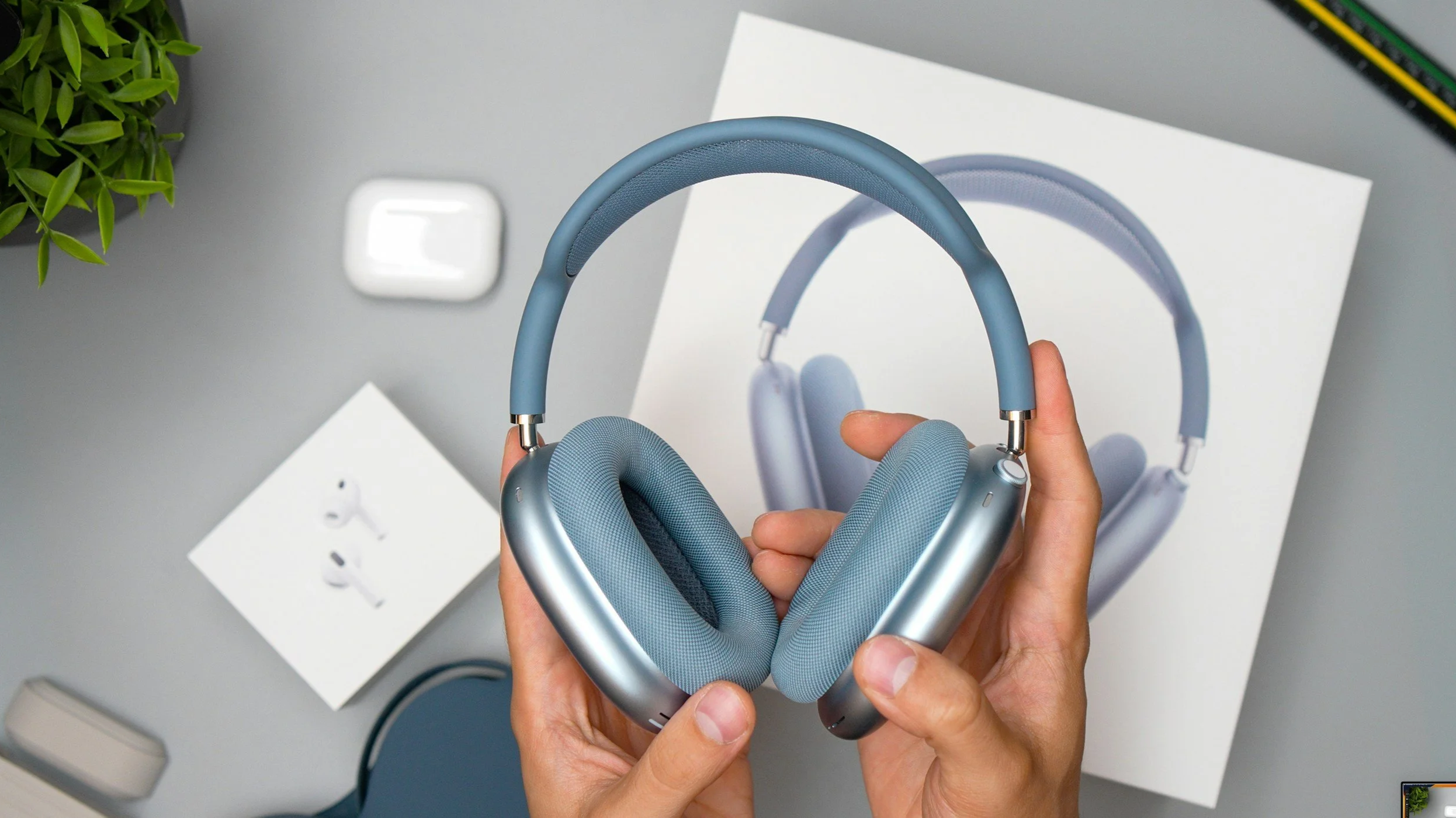 Blue Apple AirPods Max 2 held over a pair of white AirPods Pro 3