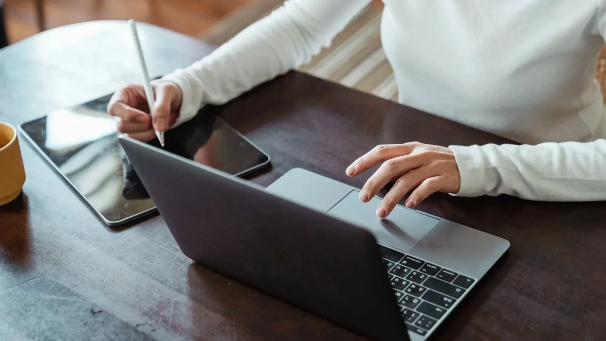 Woman using a laptop while writing on a tablet with a stylus at a wooden desk