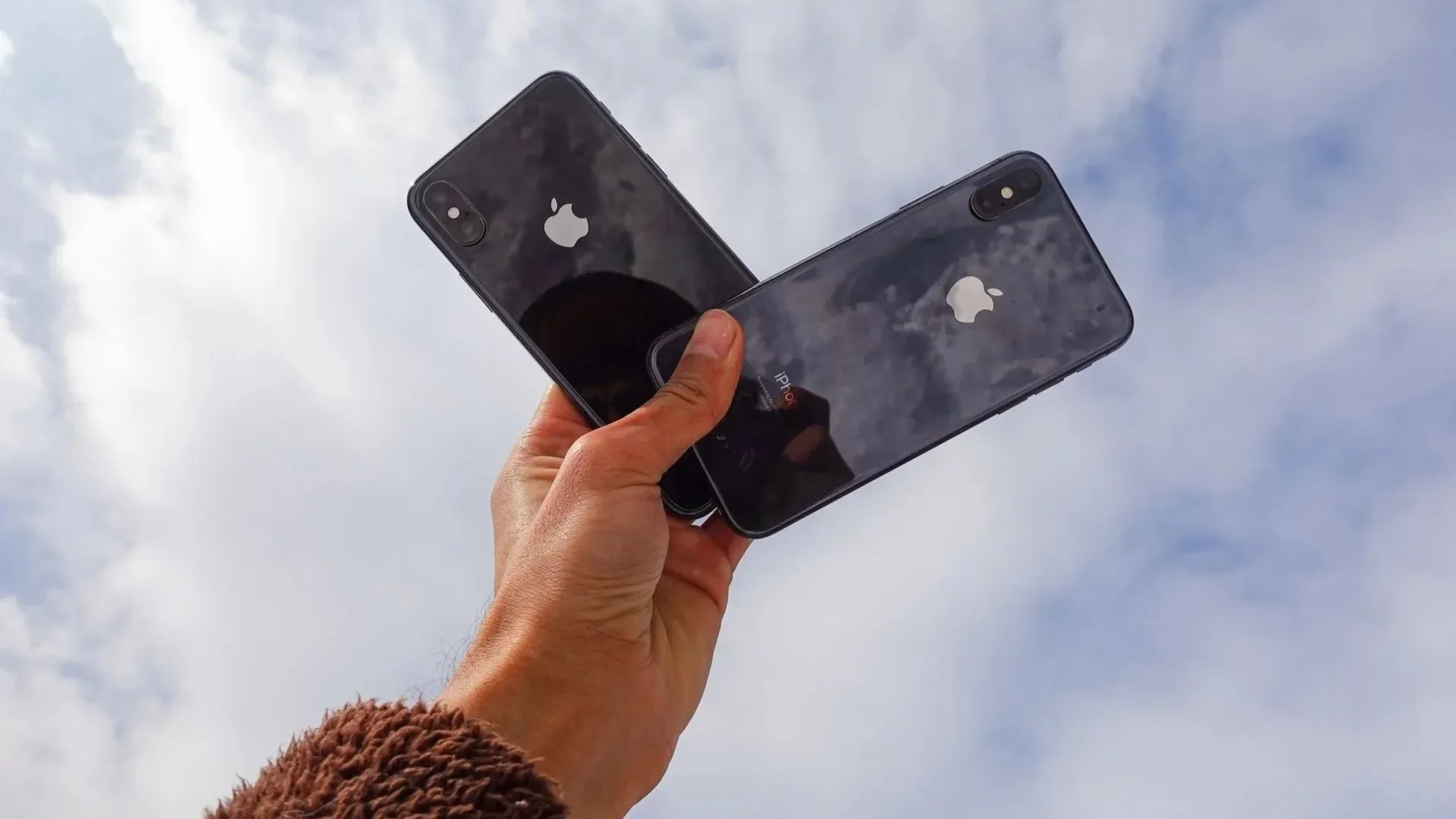 Hand holding two black iPhones up against a cloudy sky