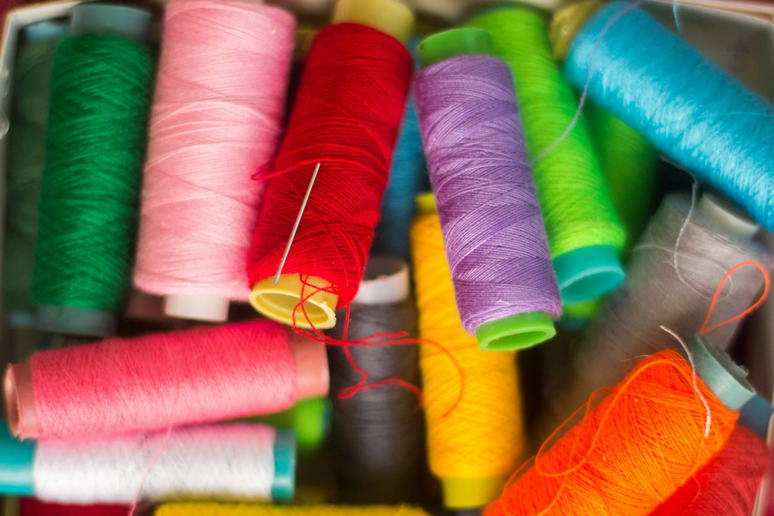 Close-up of multiple colorful sewing thread spools including pink, red, purple, green, blue, yellow, and orange, with some threads loose and tangled.