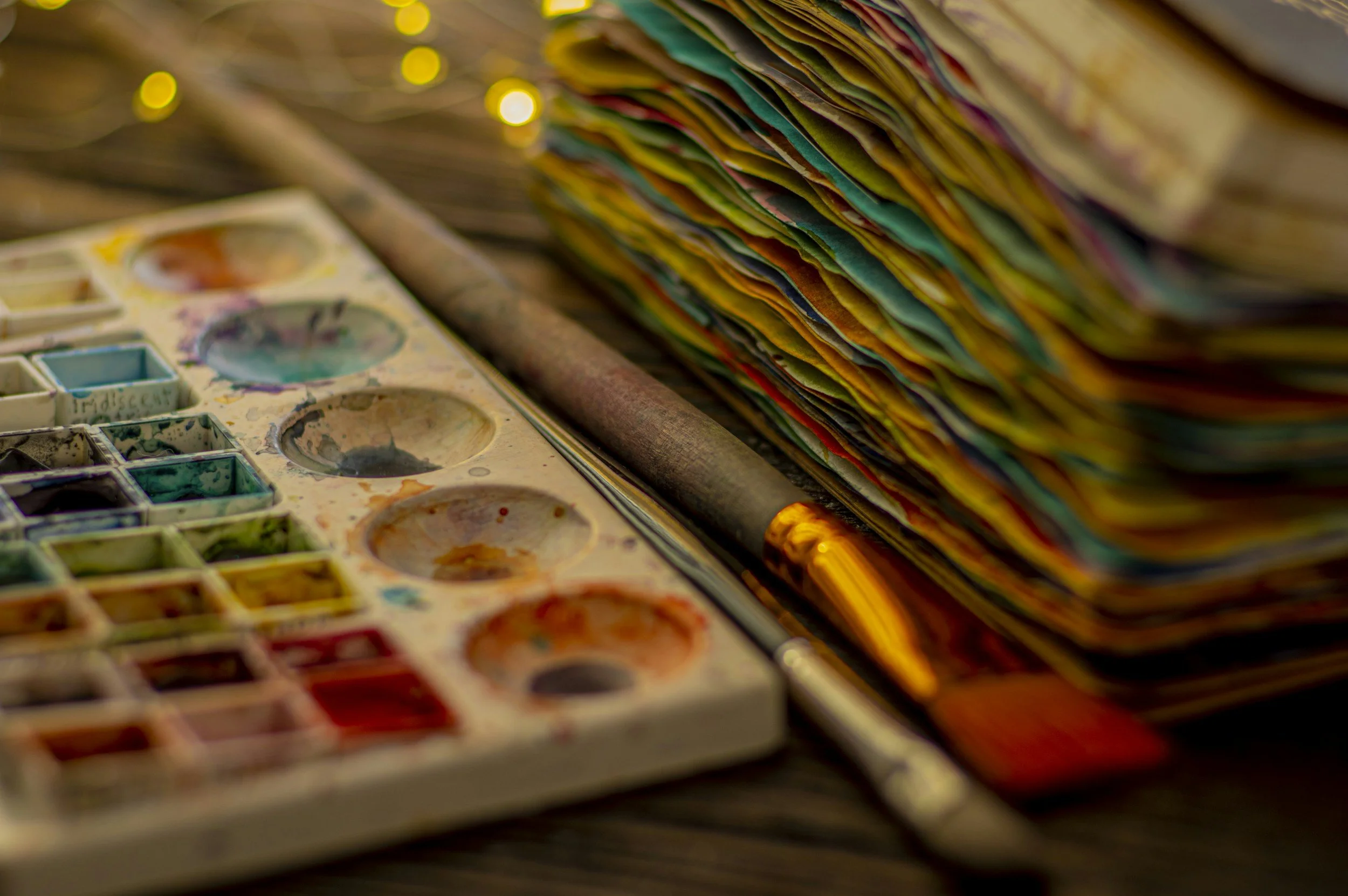 A watercolor paint palette with various colors and a stack of colorful paper or fabric behind it.