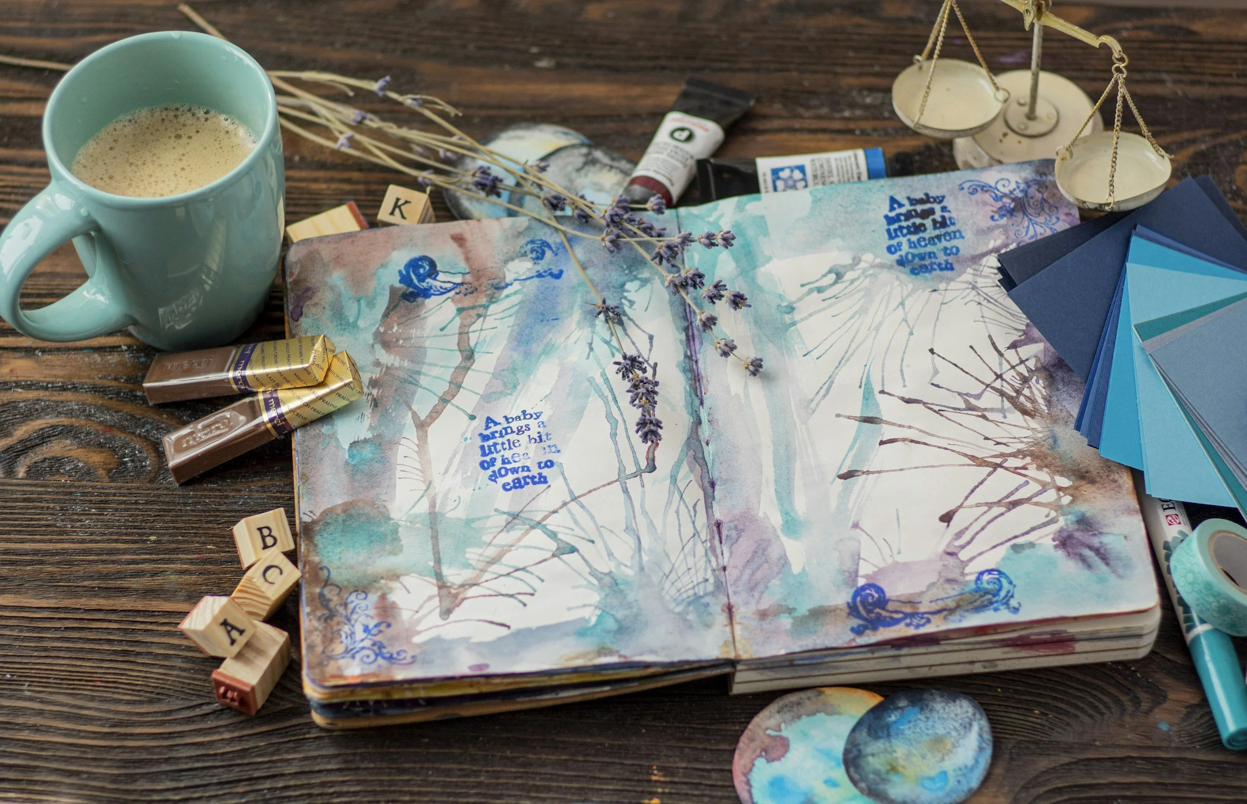 Open watercolor journal with painted trees and a bird, surrounded by lavender sprigs, paint tubes, a mug of frothy coffee, assorted blue color swatches, wooden alphabet blocks, and a small balance scale on a wooden surface.