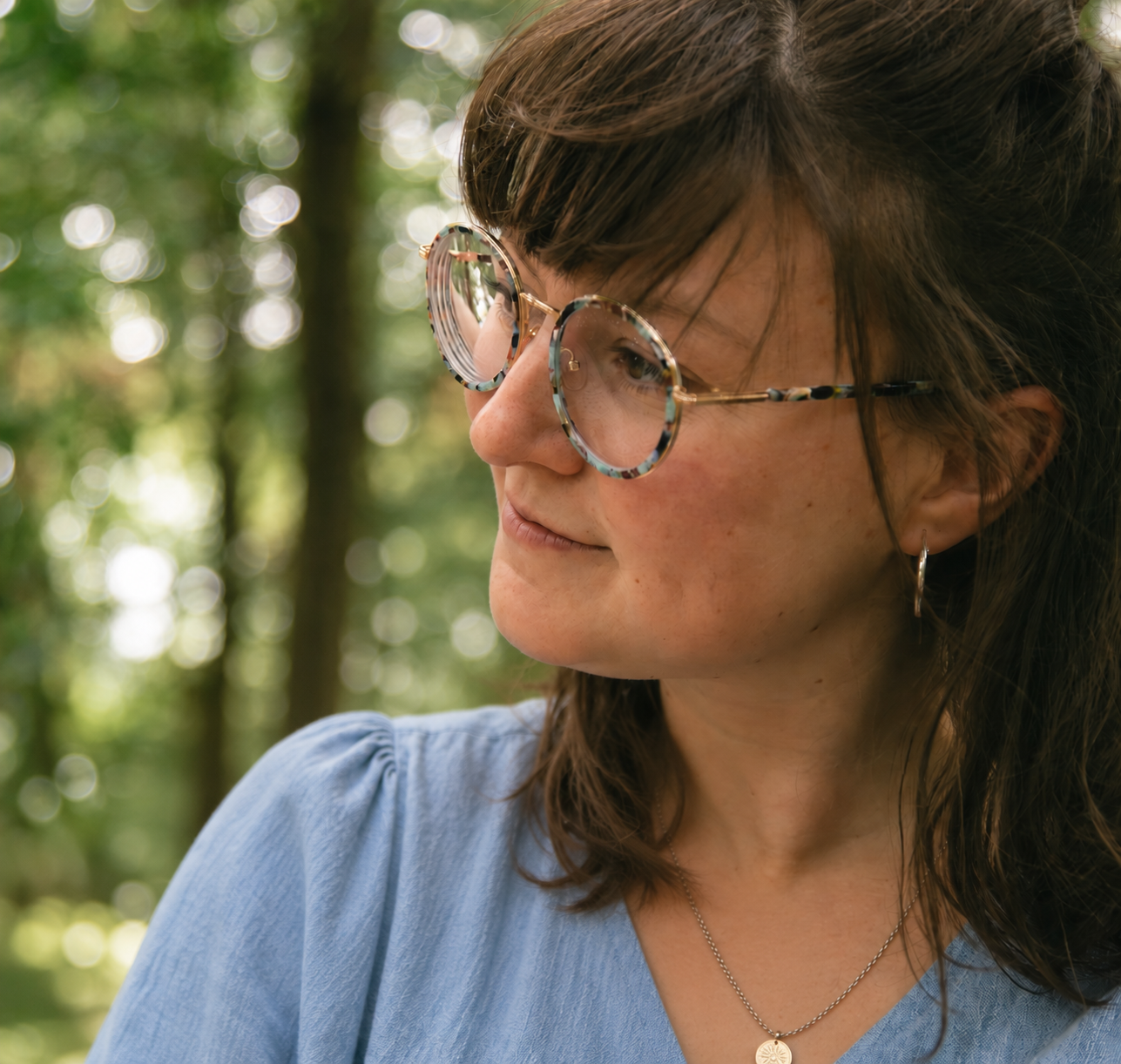 Close-up of a woman with brown hair wearing glasses, earrings, a necklace, and a light blue top, outdoors with blurred green trees in the background.