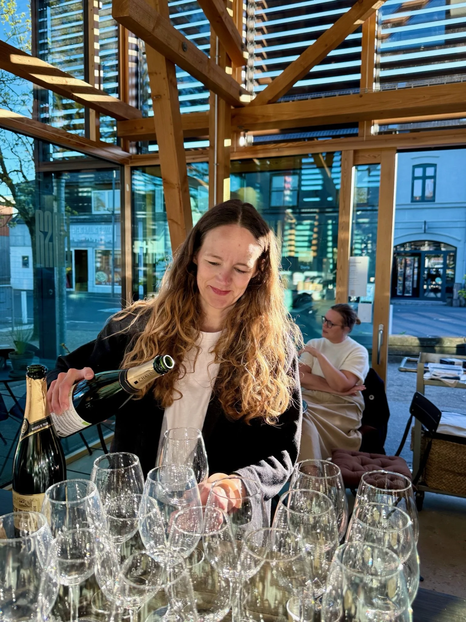 En kvinde med langt, bølget, brunt hår, der holder en champagneflaske, sidder ved et bord med flere tomme champagneglas, i en restaurant med store vinduer og træindretning, hvor to andre personer er synlige i baggrunden.