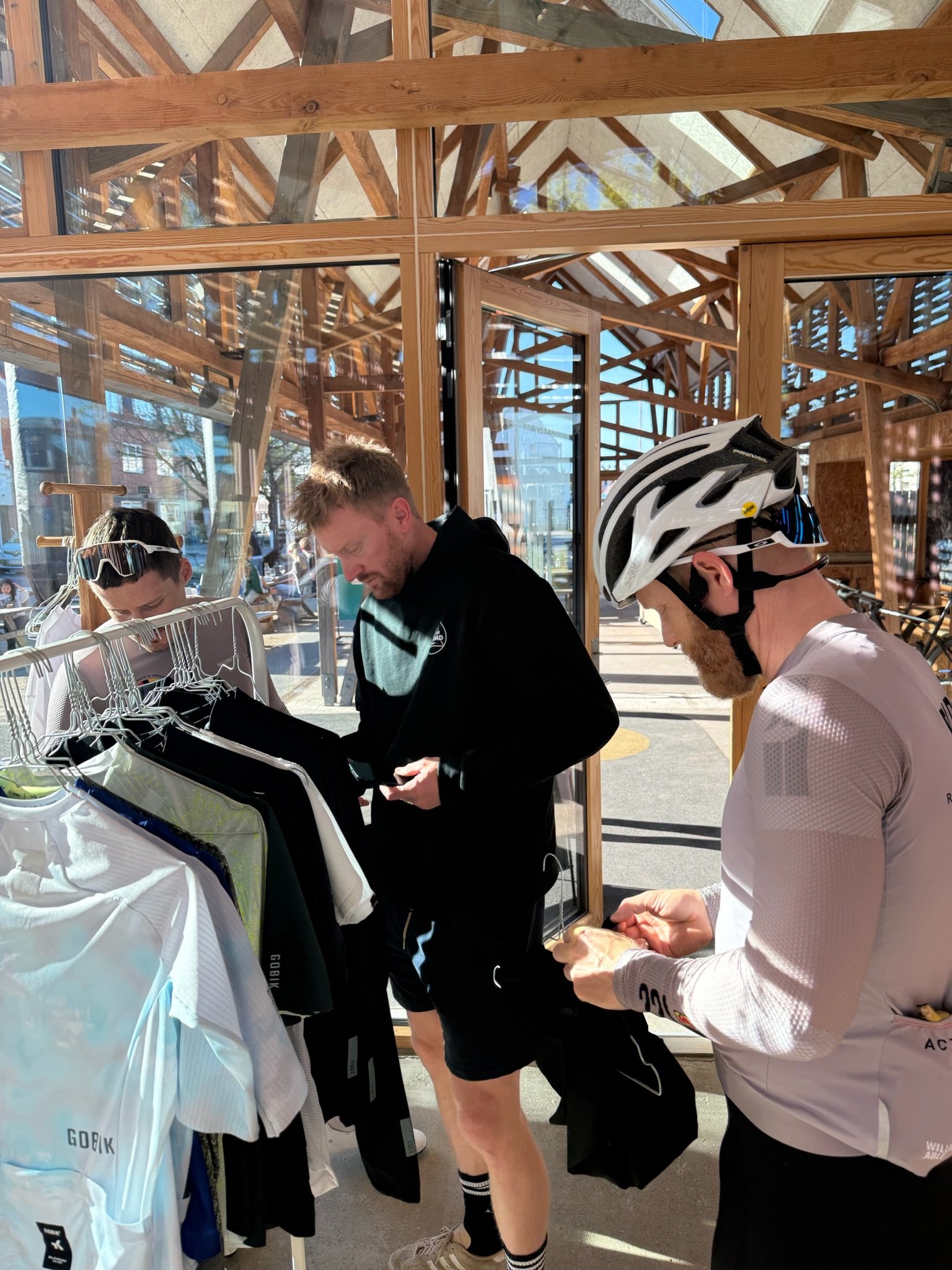 Tre mænd, der ser ud til at være deltagere i en cykelbegivenhed, står under en træinspireret shelter og gennemgår deres ting. En af dem har cykelhjelm, og de står foran en tøjholder med cykeltrøjer.