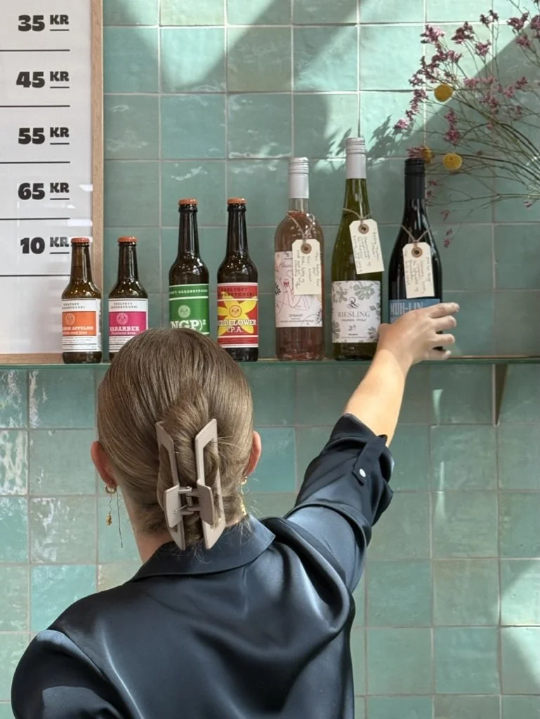 En person med enkelt hår, der har en stor hårklemme, når ud til en hylde med flasker på, i en butik eller bar. Baggrunden er grønne fliser, og der er en prismærkeskilt til venstre.