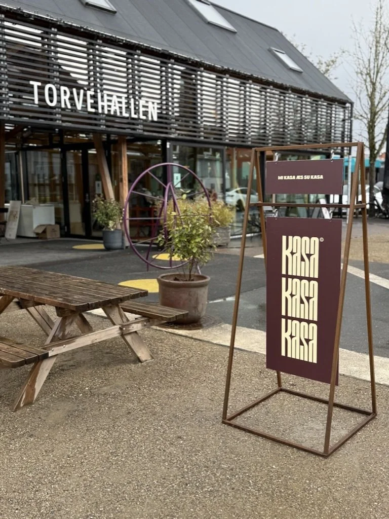 Udenfor en bygning med skiltet 'TORVEHALLEN'. Der står en bord/bænke-udendørs-opsætning, en plante i stor krukke og en reklameskilt med teksten 'KASA KASA KASA'.