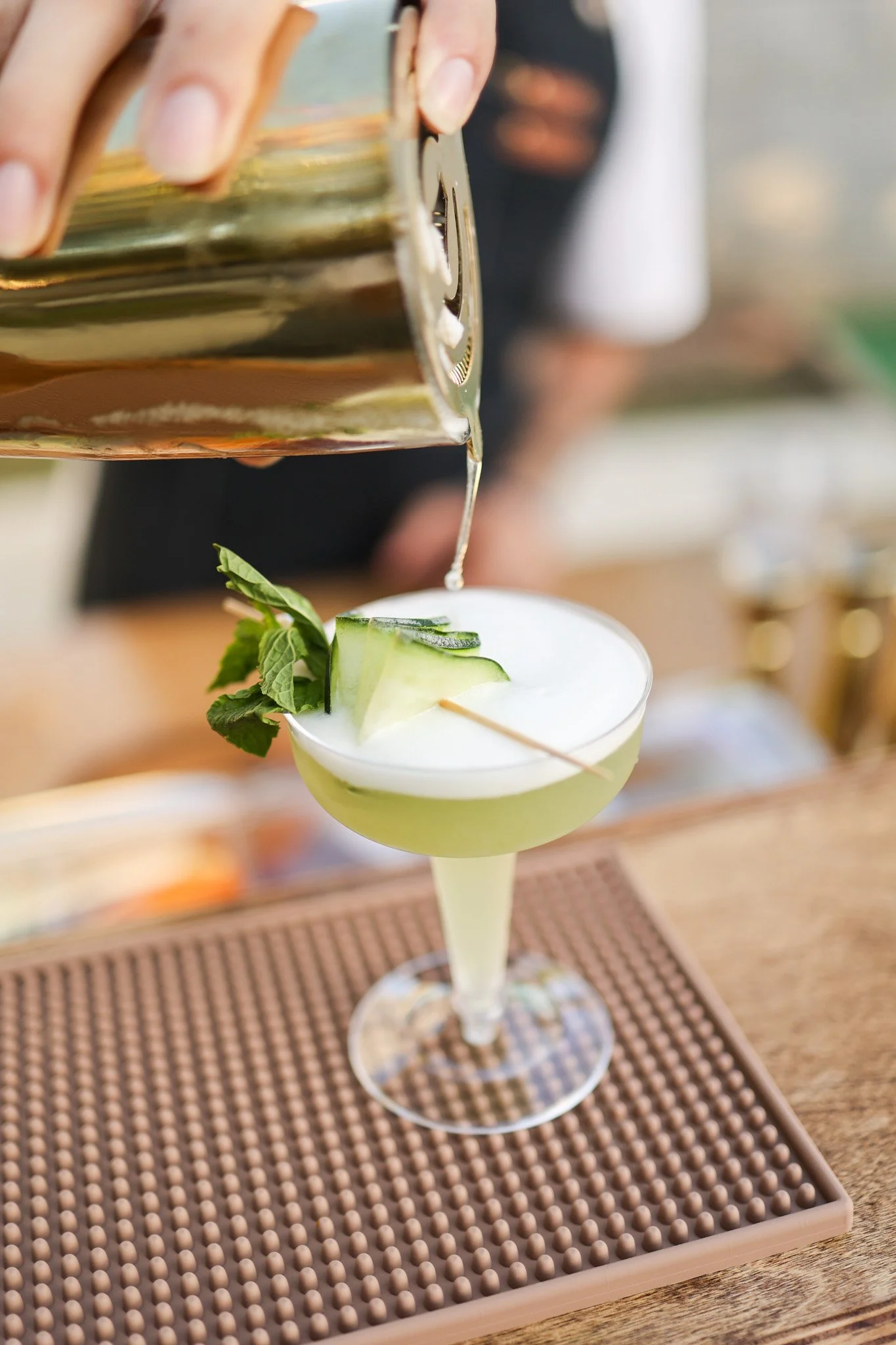 A cocktail glass filled with a light green drink, garnished with cucumber slices and mint leaves, with a person pouring a clear liquid into it.