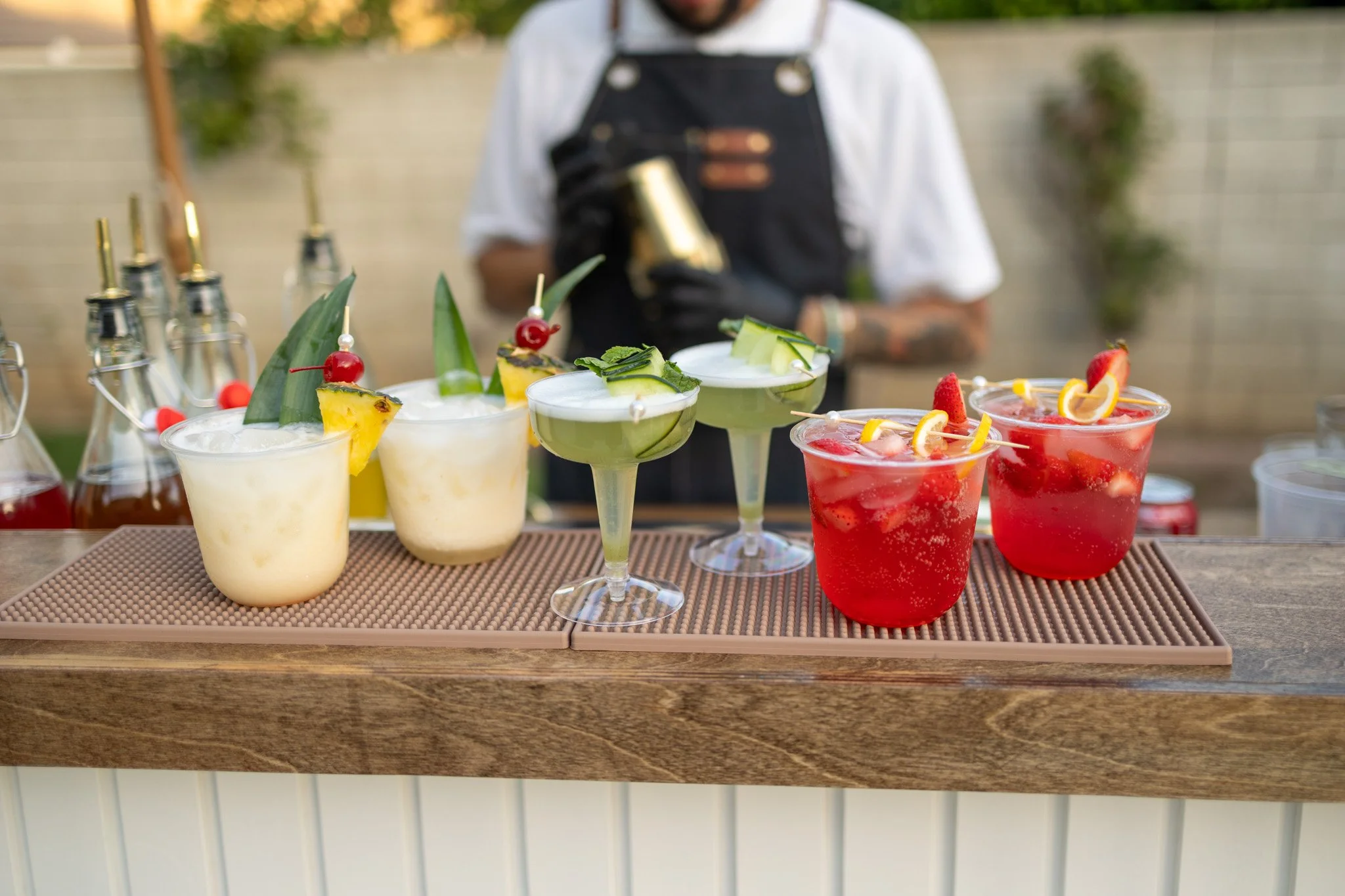 Colorful cocktails on a bar countertop with a blurred bartender in the background.