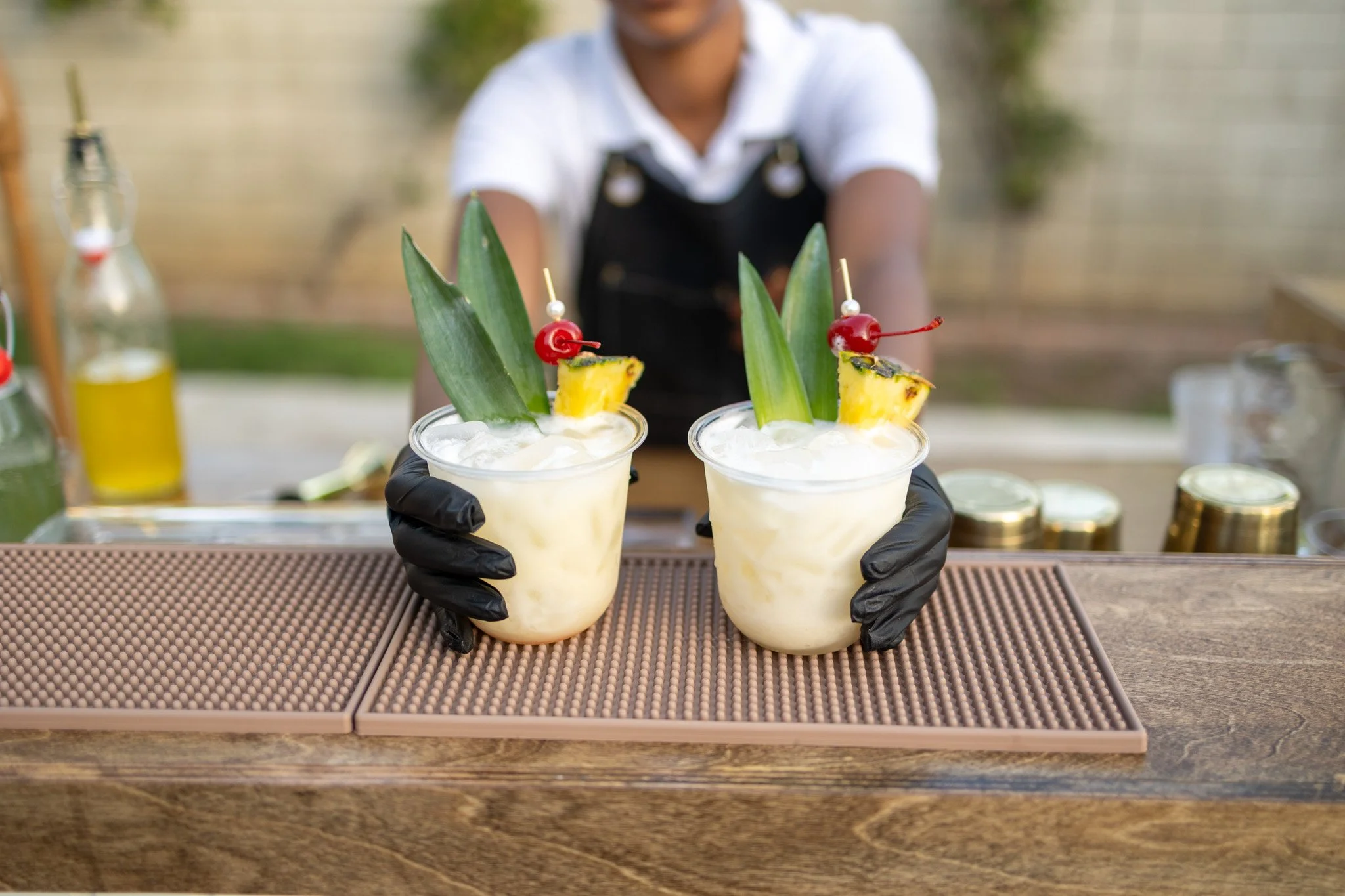 Person wearing black gloves serving two tropical drinks topped with pineapple slices, maraschino cherries on skewers, pineapple leaves, and coconut flakes, on a brown tray at outdoor setting.