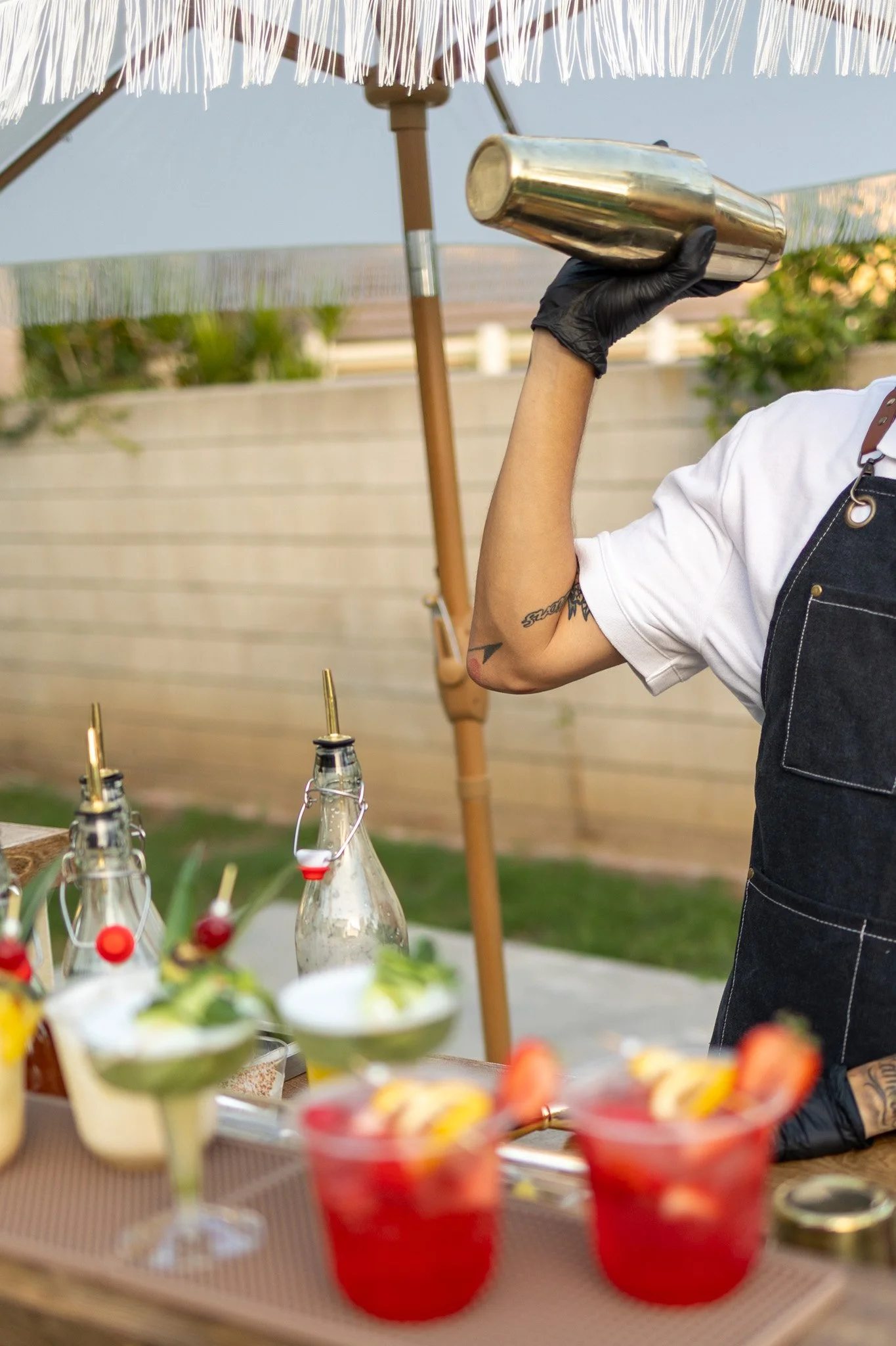 A person holding a gold cocktail shaker, wearing black gloves and a black apron, at an outdoor gathering with a table of drinks and snacks, under a large umbrella.