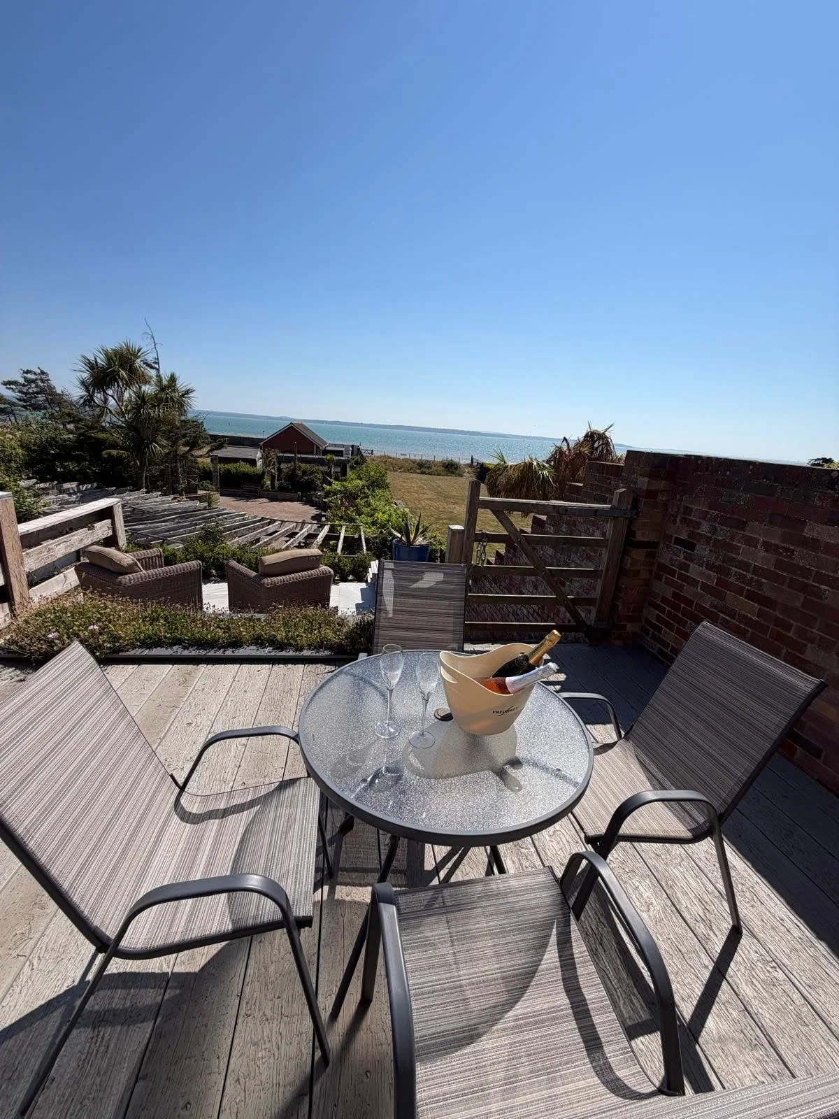 Outdoor patio with a glass table set with two wine glasses, a bucket with a wine bottle, and four chairs, overlooking a garden, wooden steps, and a view of the ocean under a clear blue sky.