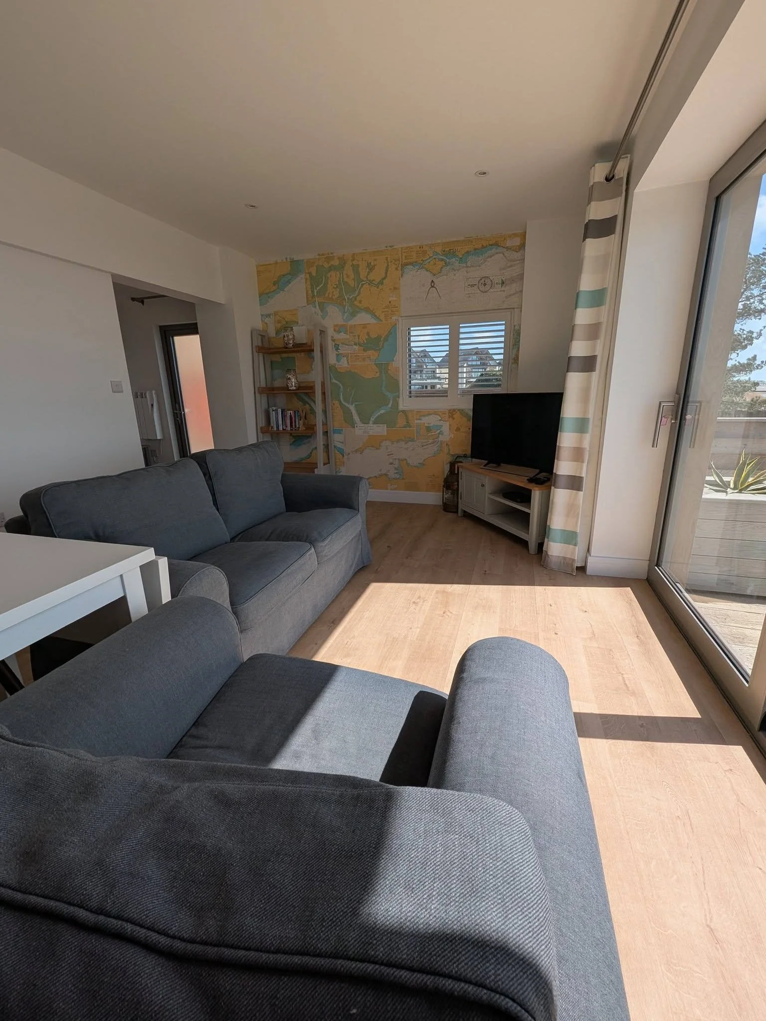 Living room with a gray sofa, a wooden TV stand with a flat-screen TV, a bookshelf, large windows with striped curtains, and a wall with a colorful city map mural.