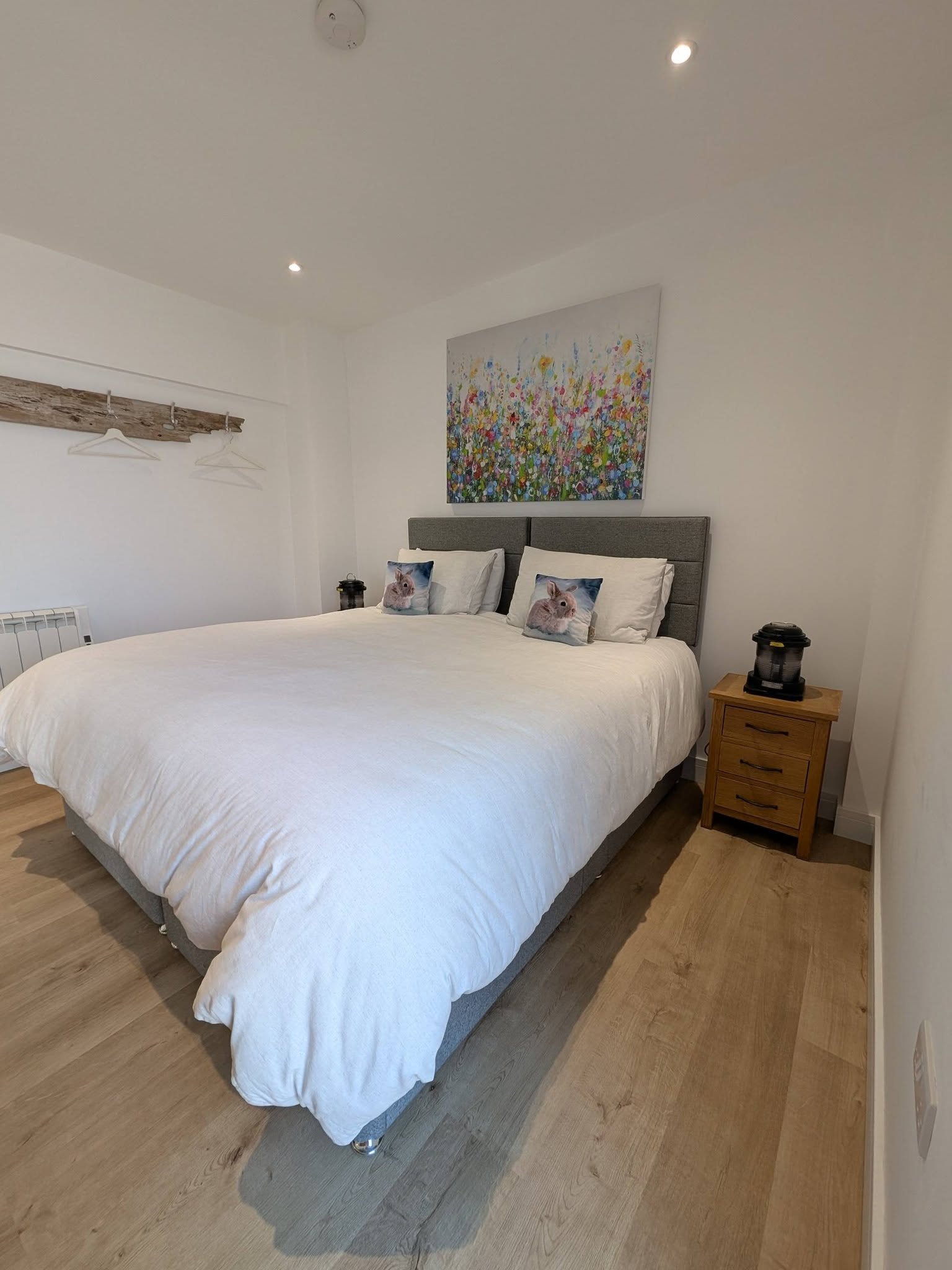 A bedroom with white bedding, two pillows with bunny prints, a gray upholstered headboard, a wooden nightstand with a black lantern-style light, a landscape painting of colorful flowers above the bed, a wooden wall rack with hangers, and hardwood floors.