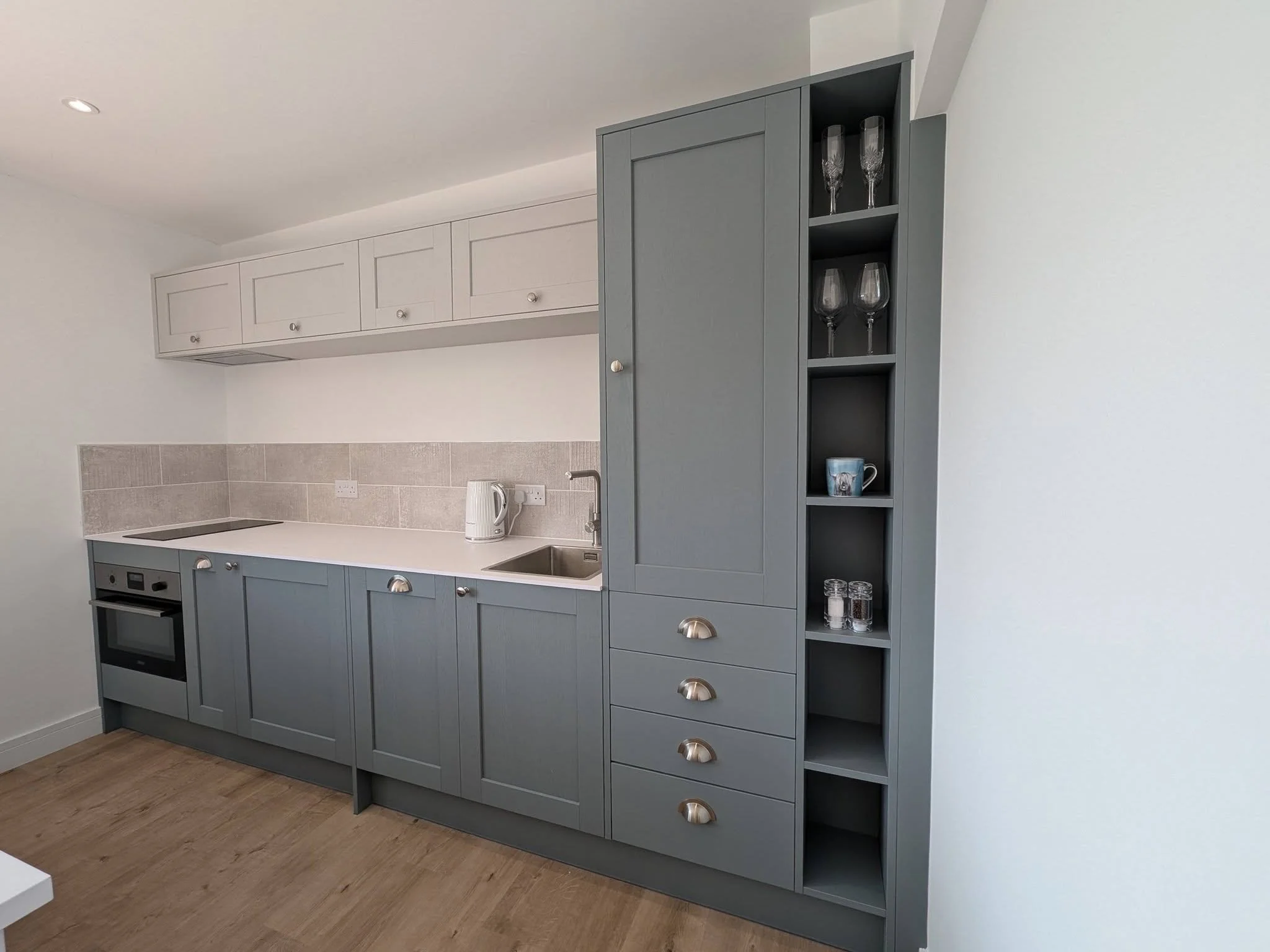 A kitchen with blue cabinets, a white countertop, a built-in oven, and a gray cabinet with glassware and a mug.