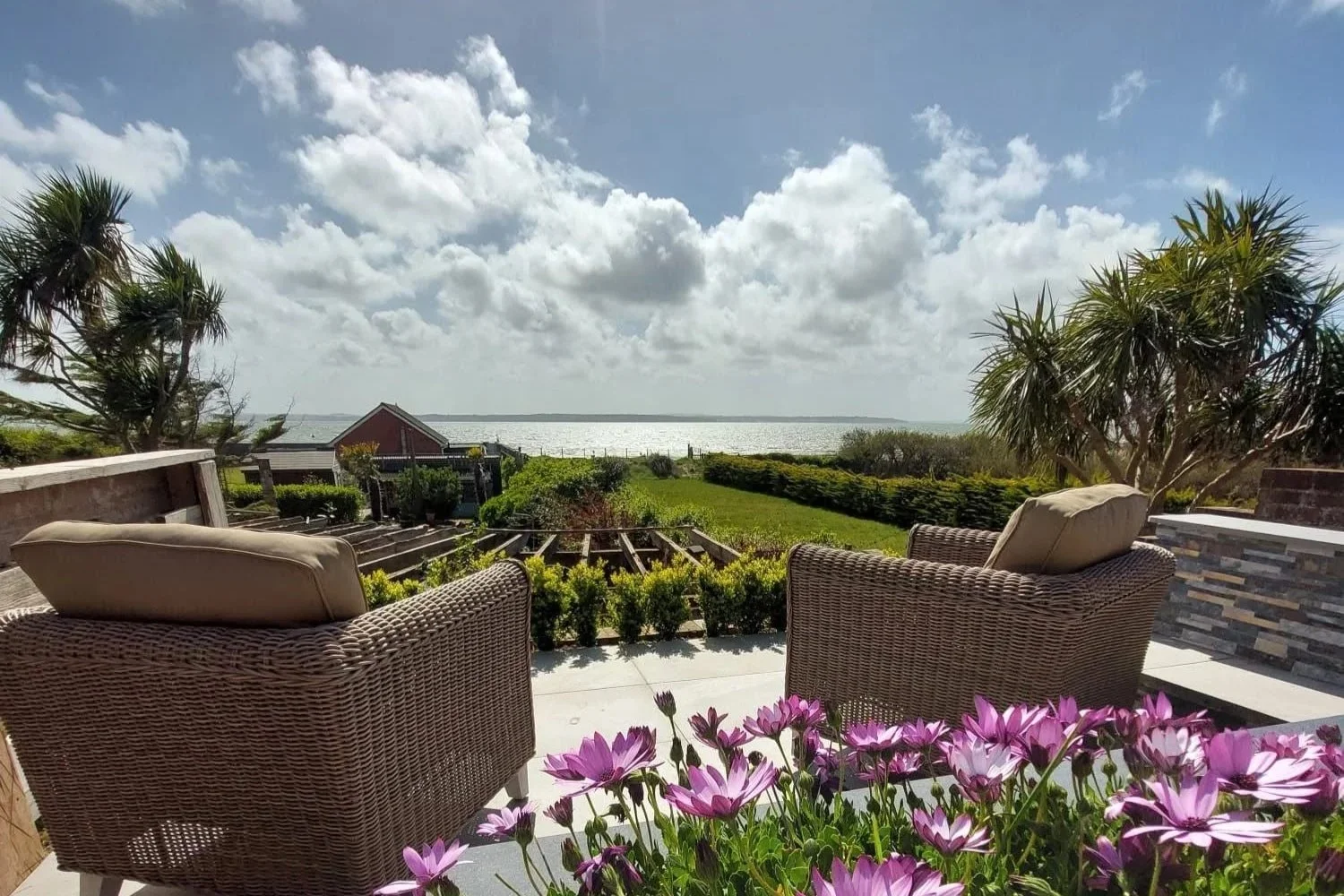 A view of a backyard patio with wicker chairs, pink flowers in the foreground, lush greenery, and a body of water in the distance under a partly cloudy sky.
