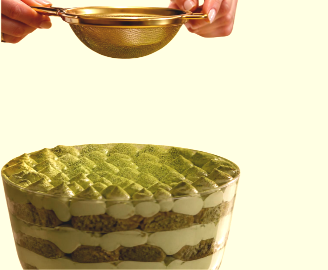 A person is using a gold strainer over a layered matcha cake with green matcha topping and white cream filling, set against a light background.