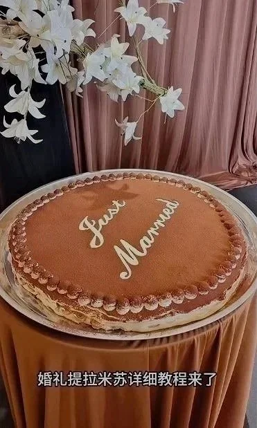 A large round tiramisu with a smooth chocolate surface and the words "Just Married" written on it, placed on a table with draped fabric and white flowers in the background.