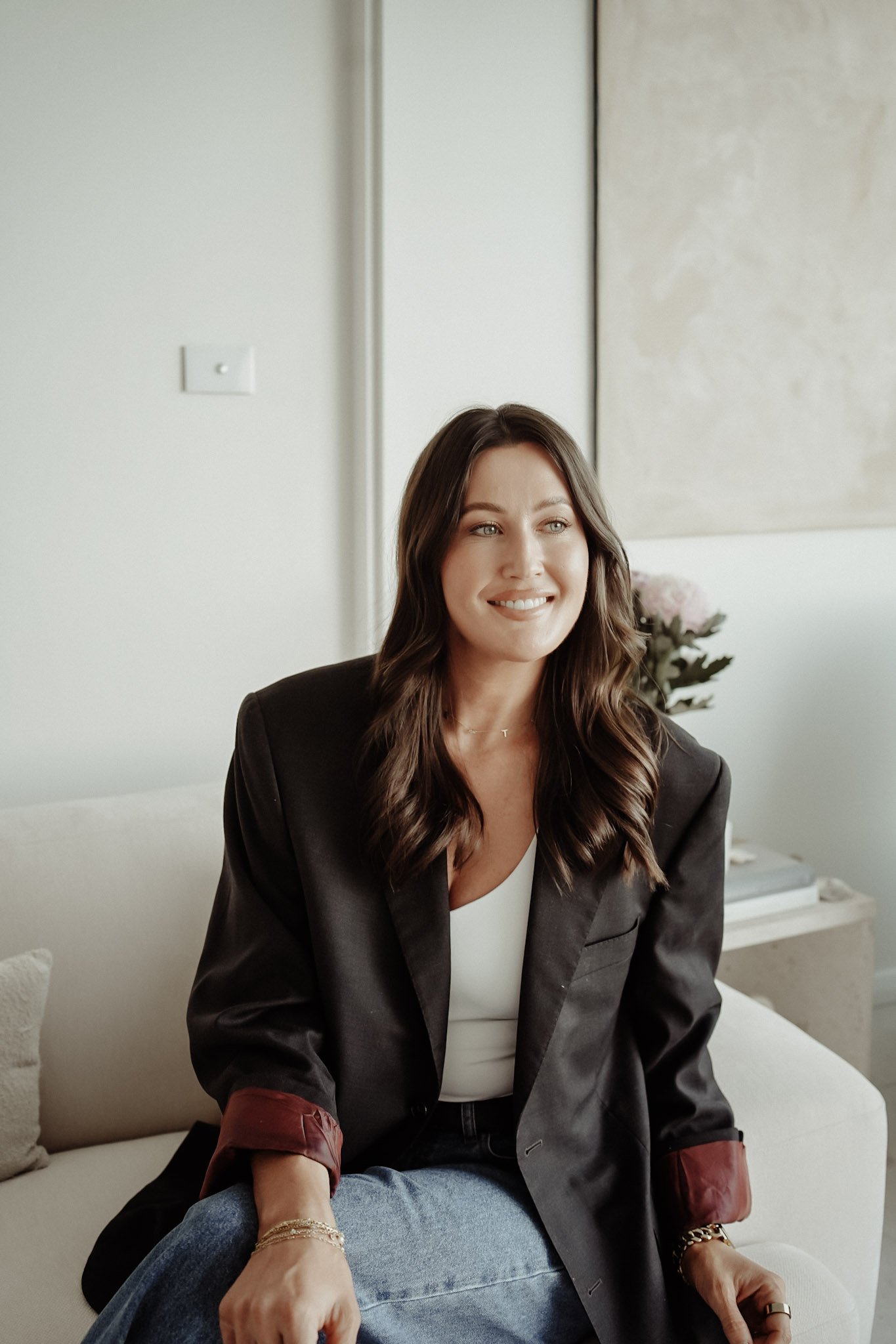 A woman with long brown hair smiling, sitting on a sofa in a room with light-colored walls and a large artwork, wearing a dark blazer, white top, and blue jeans, with a person beside her wearing a gold bracelet and ring.