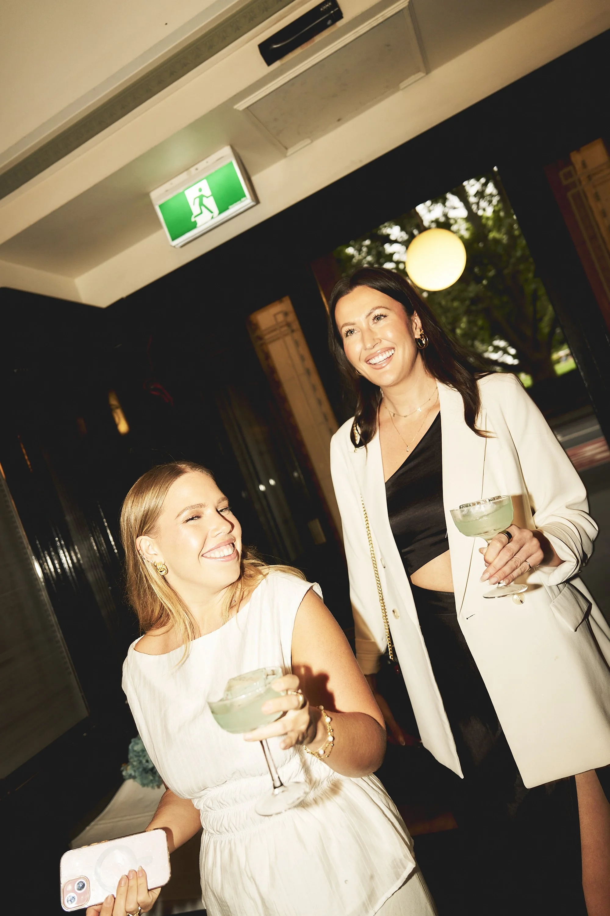 Two women smiling and holding drinks at a social event, with one woman taking a selfie. One woman is wearing a white dress, and the other is wearing a white blazer over black. The setting appears to be indoors with a window showing trees outside and 