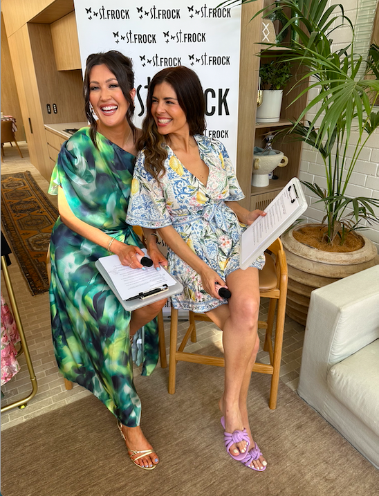 Two women sitting on chairs indoors, smiling and holding clipboards, with a branded backdrop behind them that reads 'st.frock'. One woman is standing, wearing a green and blue patterned dress, and the other woman is seated, wearing a floral dress and