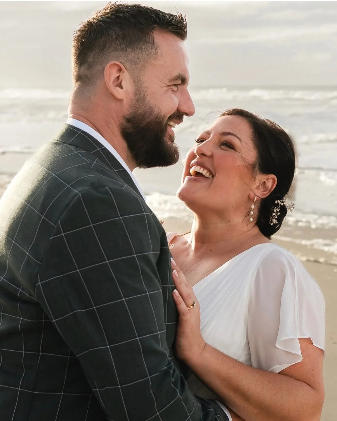 A couple dressed in wedding attire sharing a joyful moment on the beach with the ocean in the background.