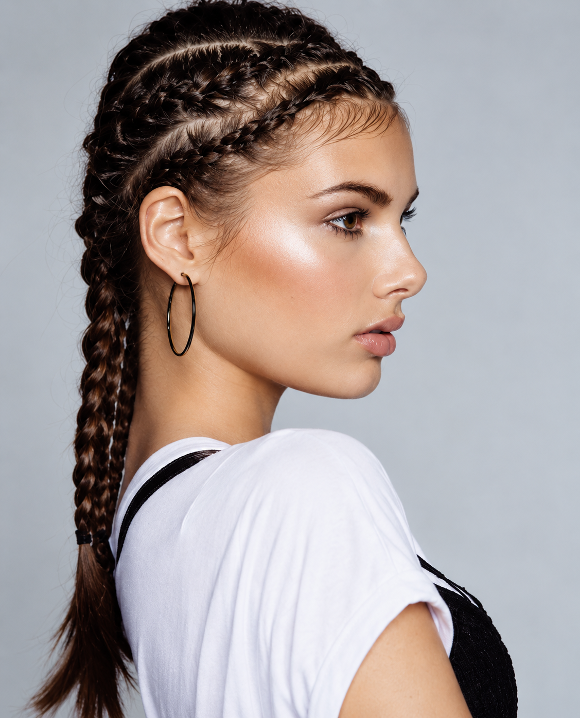Profile of a young woman with braided hair, hoop earrings, and natural makeup, wearing a white shirt with black straps, against a plain background.