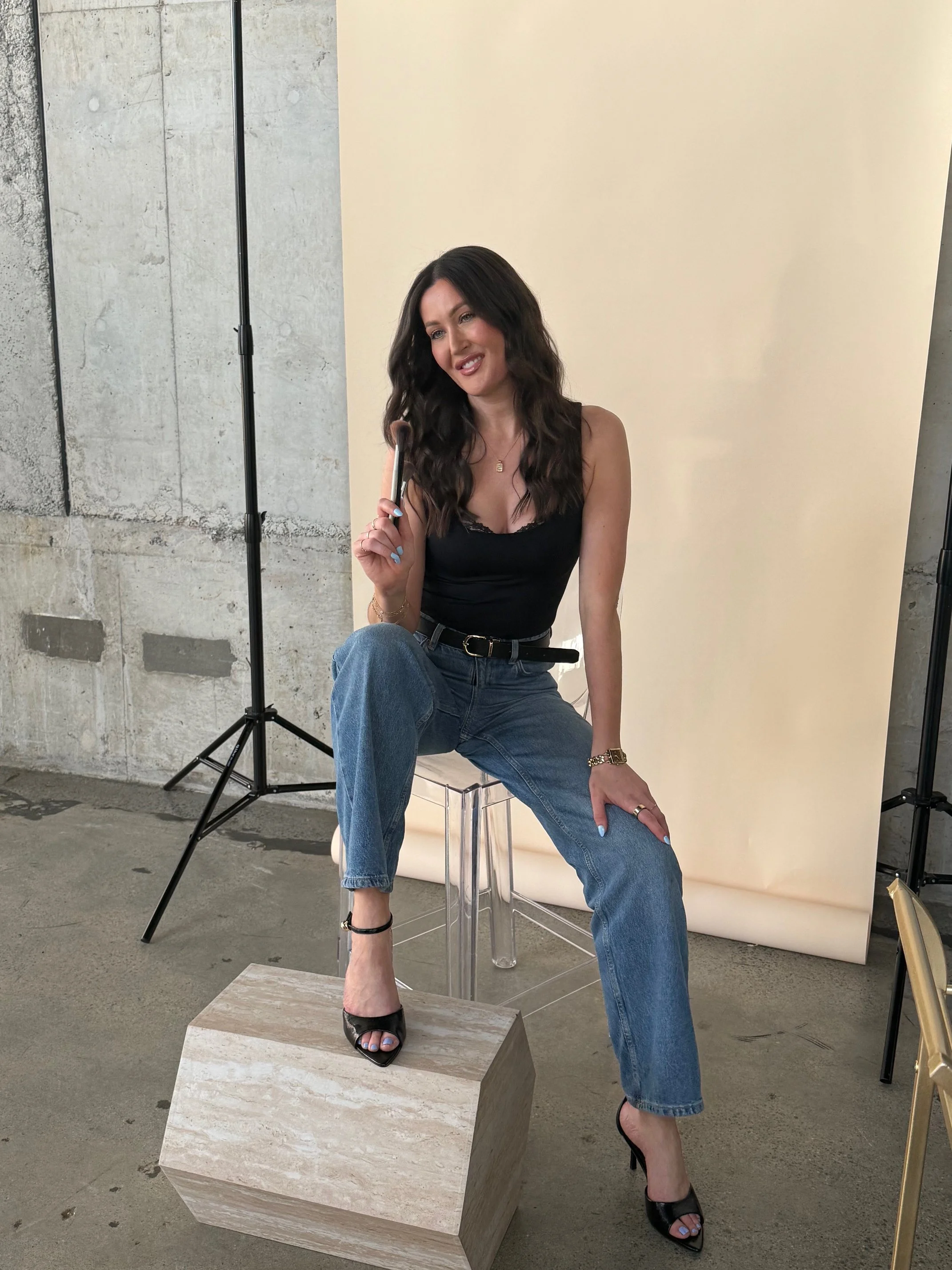 A woman with long dark hair sitting on a transparent chair, holding a makeup brush, wearing a black top and blue jeans, with one foot elevated on a small beige stone block in what appears to be a studio with a concrete wall and a beige backdrop.