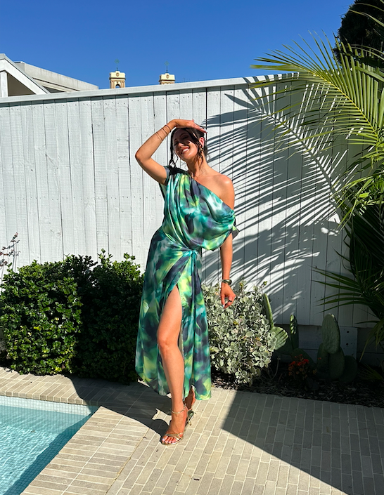 Woman in colorful dress standing by a poolside, smiling with one hand on her head, surrounded by plants and a white fence, sunny day.