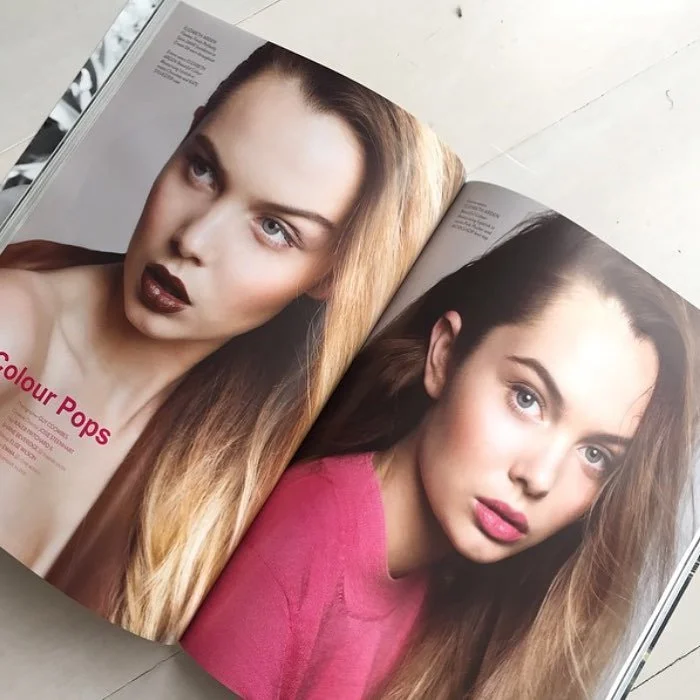 Open magazine showing a close-up image of a woman with long blonde hair, dark lipstick, and makeup, next to another image of a woman with long brown hair, wearing a pink top.