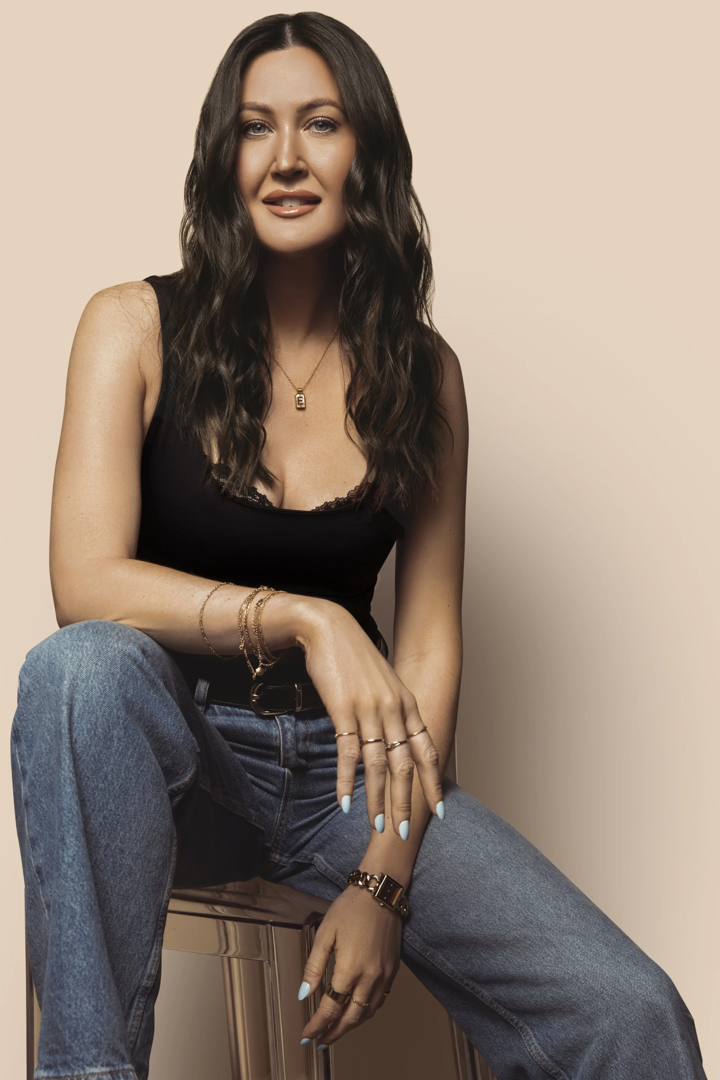 A woman with long wavy brown hair wearing a black tank top, jeans, and gold jewelry, sitting on a stool against a beige background.