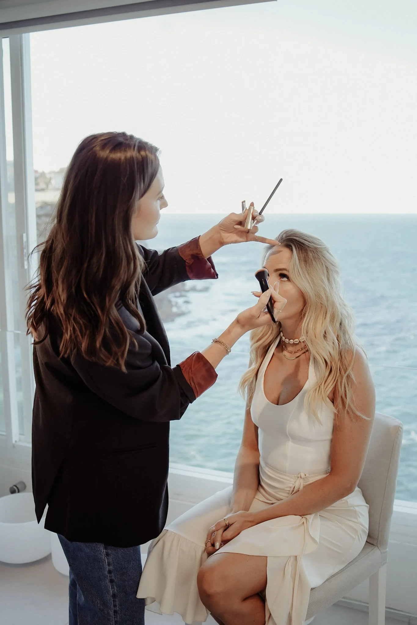 Makeup artist applying makeup to a woman with blonde hair sitting on a chair near a window overlooking the ocean.