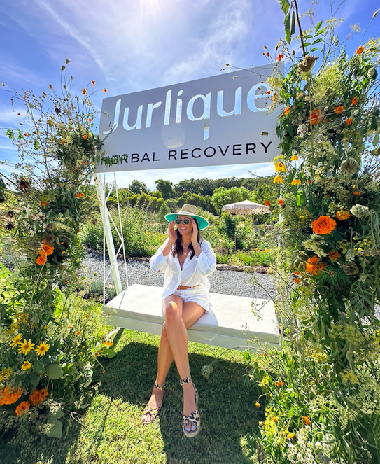 A woman sitting on a swing with a floral arch and a sign that says 'Jurlique' and 'Global Recovery' above her outdoors on a sunny day.
