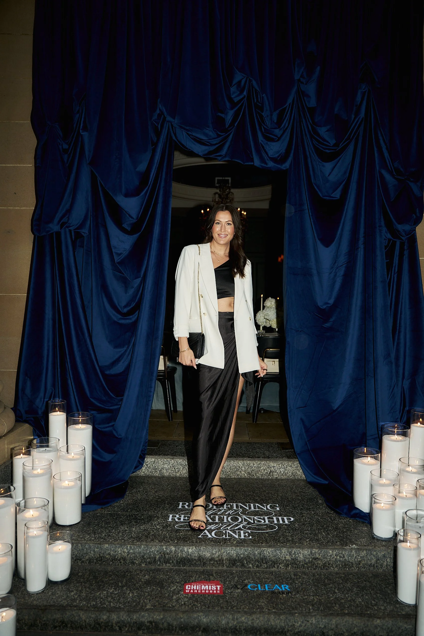 A woman walking down steps decorated with white candles in glass holders, navy blue curtains framing the entrance, and the words 'Rebuild, Reconciliation, Resilience, Love, Acne' printed on the steps.
