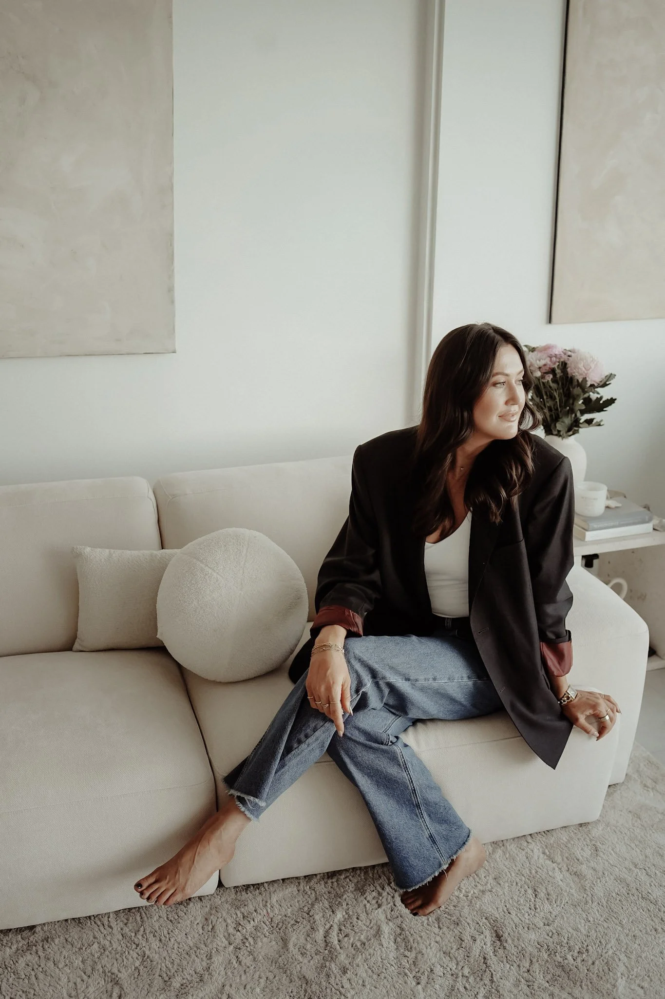 A woman with dark hair sitting on a cream-colored sofa with two decorative pillows, one round and fluffy, in a modern living room. She is looking to her right, wearing a black blazer, white top, and distressed jeans, with a bouquet of pink flowers on