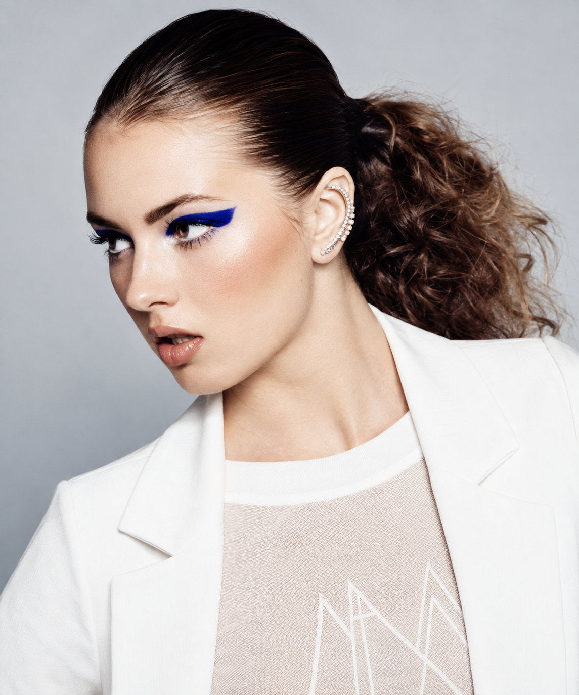 A woman with dark brown hair styled in a curly ponytail, wearing bold blue eye makeup, a white blazer, and a clear top with a geometric design, accessorized with a rhinestone ear cuff.