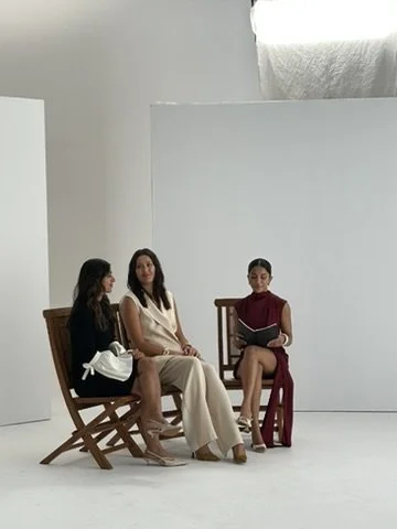 Three women sitting on chairs in a white studio, one reading a book.