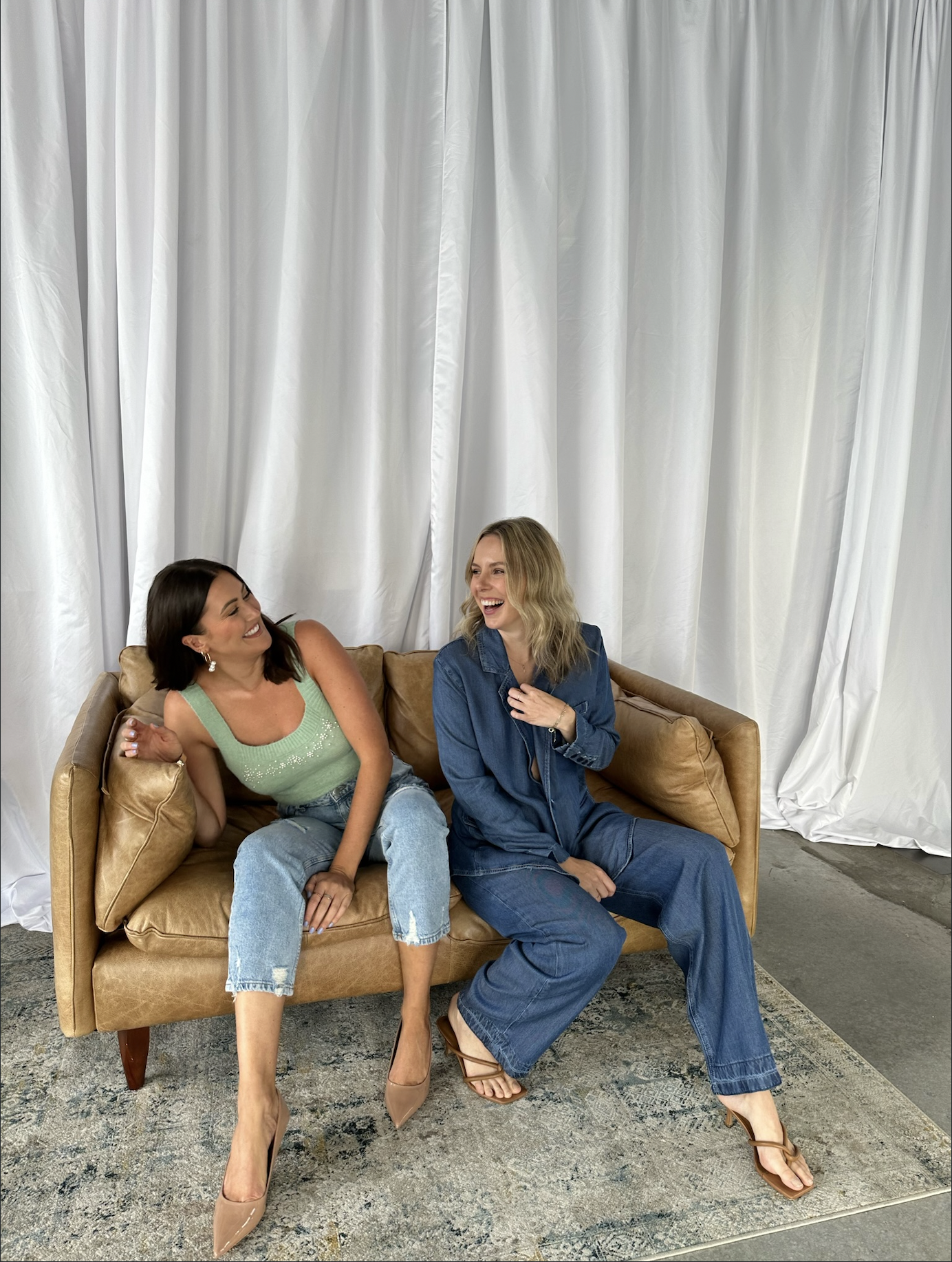 Two women laughing and sitting on a tan couch in front of a white curtain backdrop.