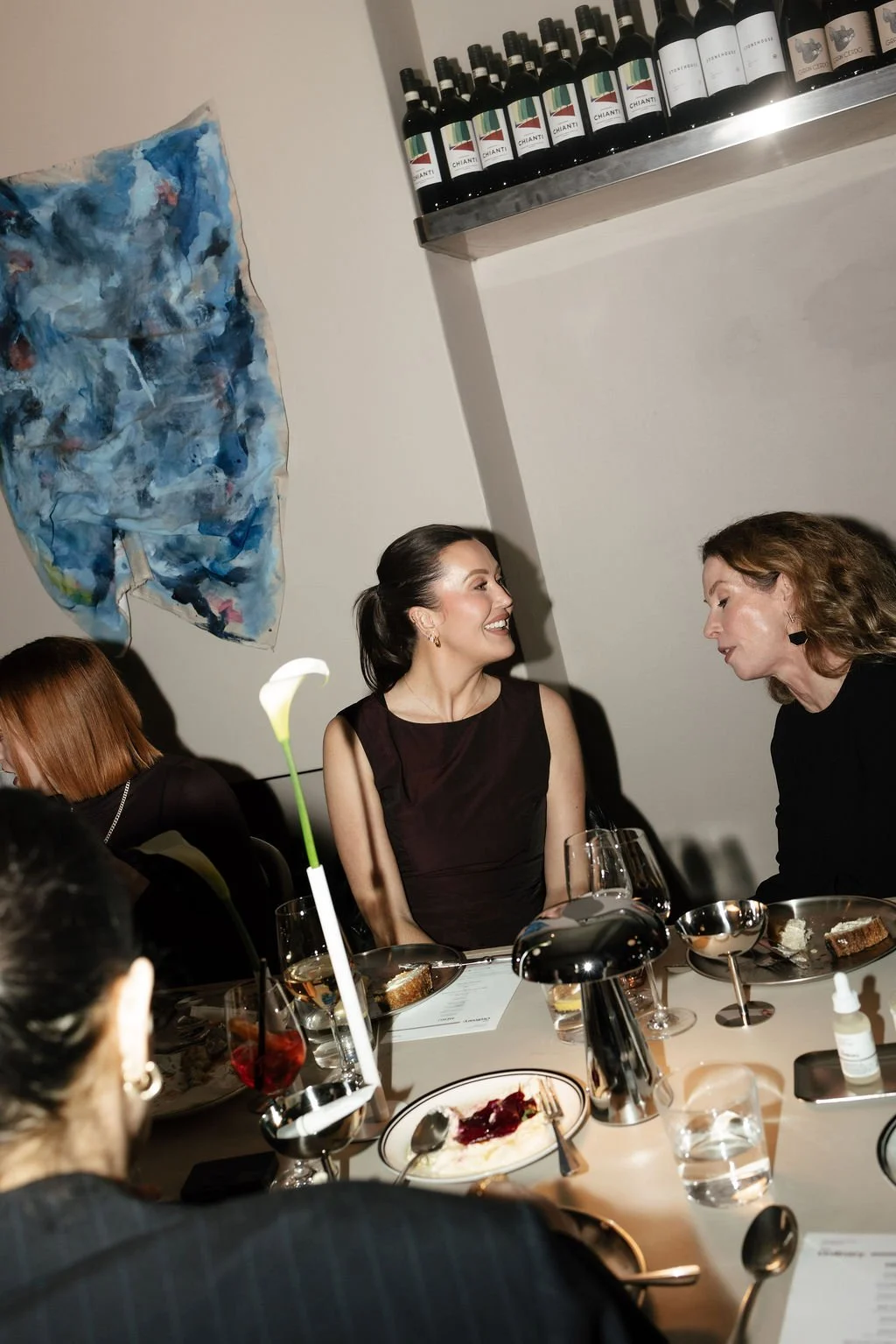 People sitting at a dinner table in a restaurant, smiling and engaging in conversation. The table has plates with food, glasses of drinks, and a flower centerpiece. There are wine bottles on a shelf against the wall.