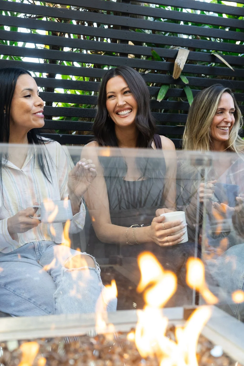 Three women sitting outside near a fire pit, holding mugs, smiling and chatting, with a wooden slat fence and green plants in the background.