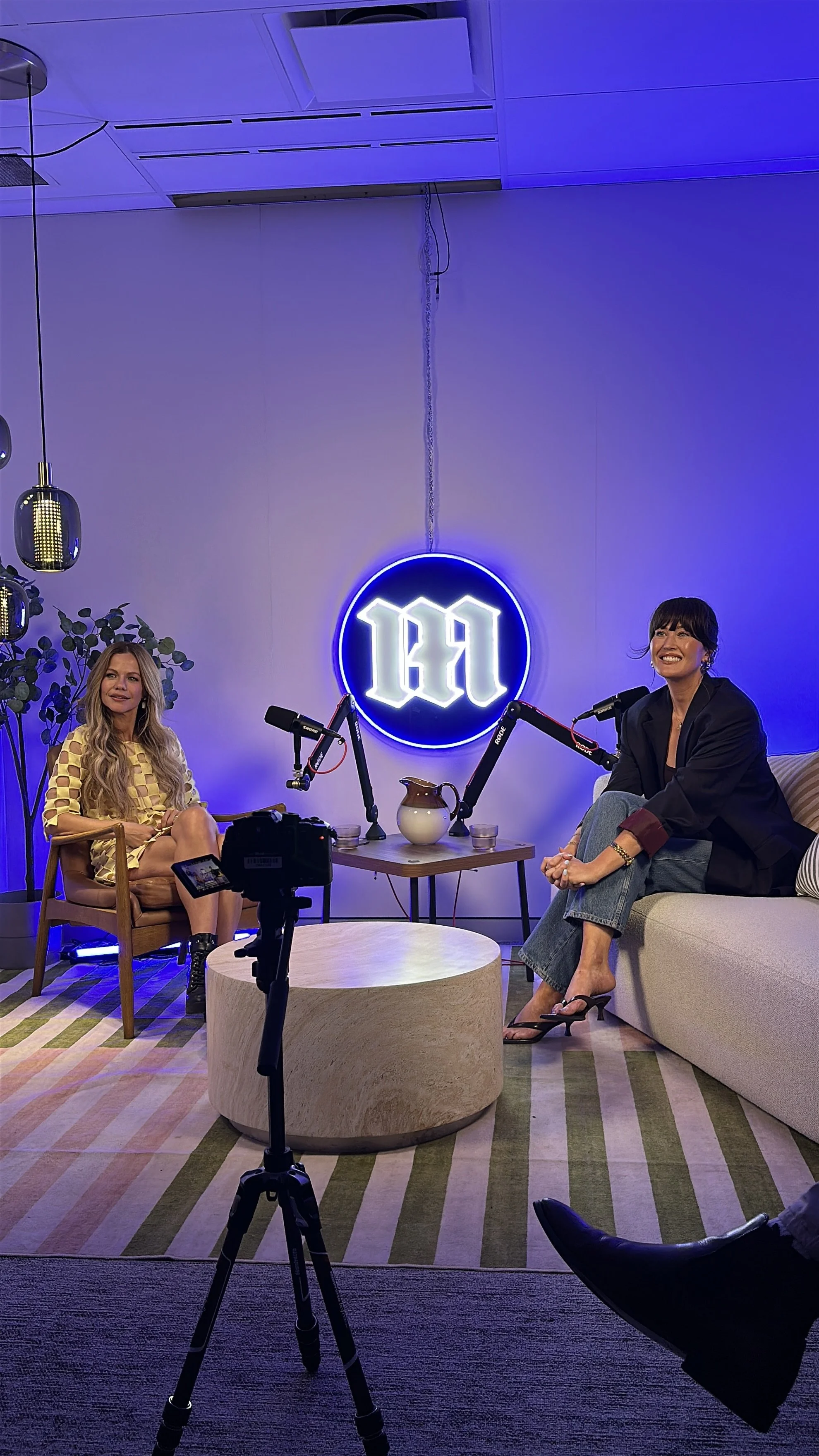 Two women sitting in a modern, stylish room with purple and blue lighting. One woman has long blond hair, wearing a yellow checkered dress, sitting in a wooden chair. The other woman has dark hair with bangs, wearing a dark blazer and jeans, sitting 