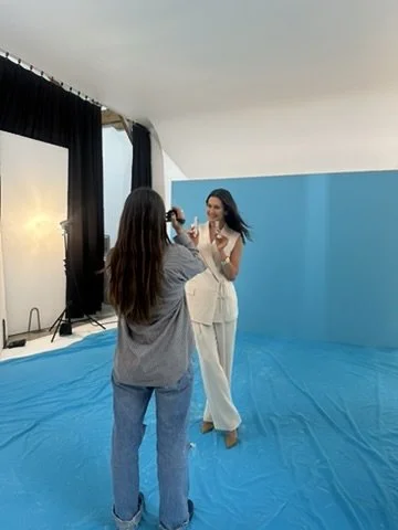 Two women in an indoor photo studio, one with long straight hair wearing a blue outfit, taking a photo of the other woman with long dark hair, wearing a white jumpsuit, standing on a blue backdrop.