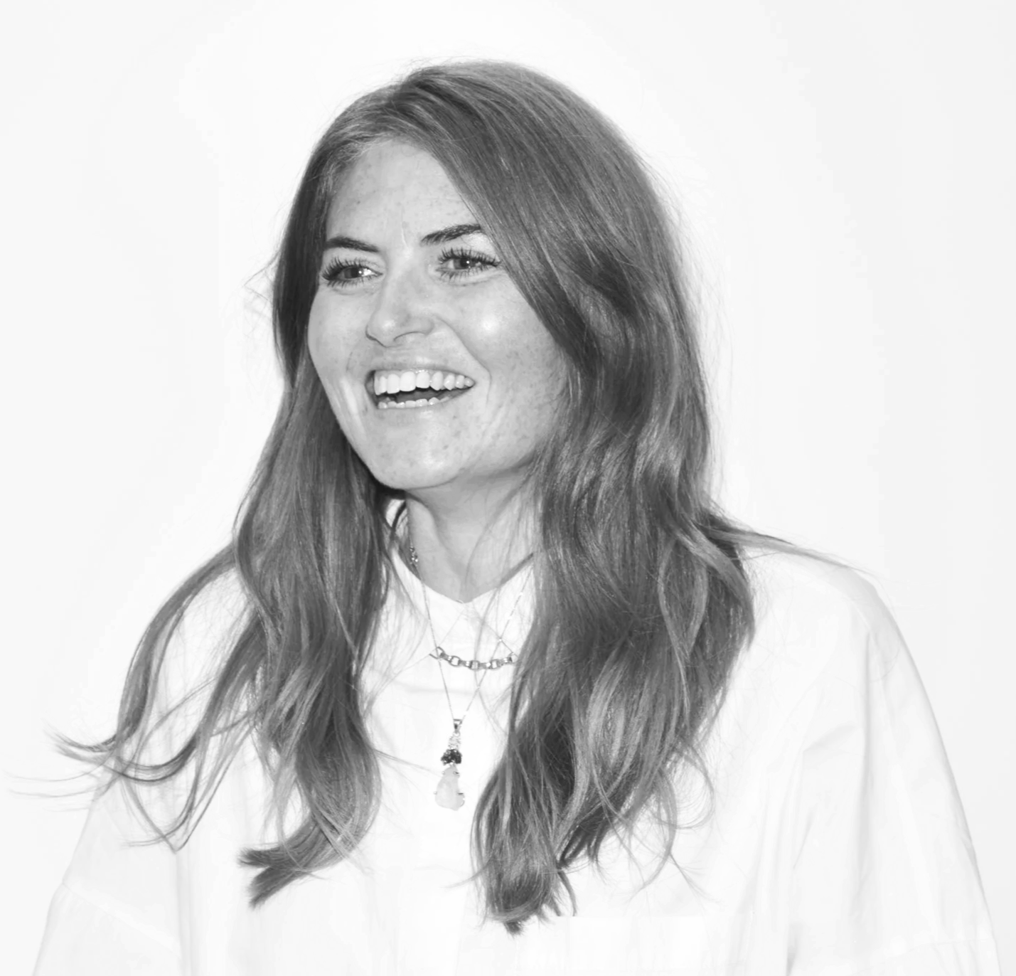 Sam Maitland in a black and white photo with long wavy hair, smiling and looking to her right, wearing a light-colored top and layered necklaces.
