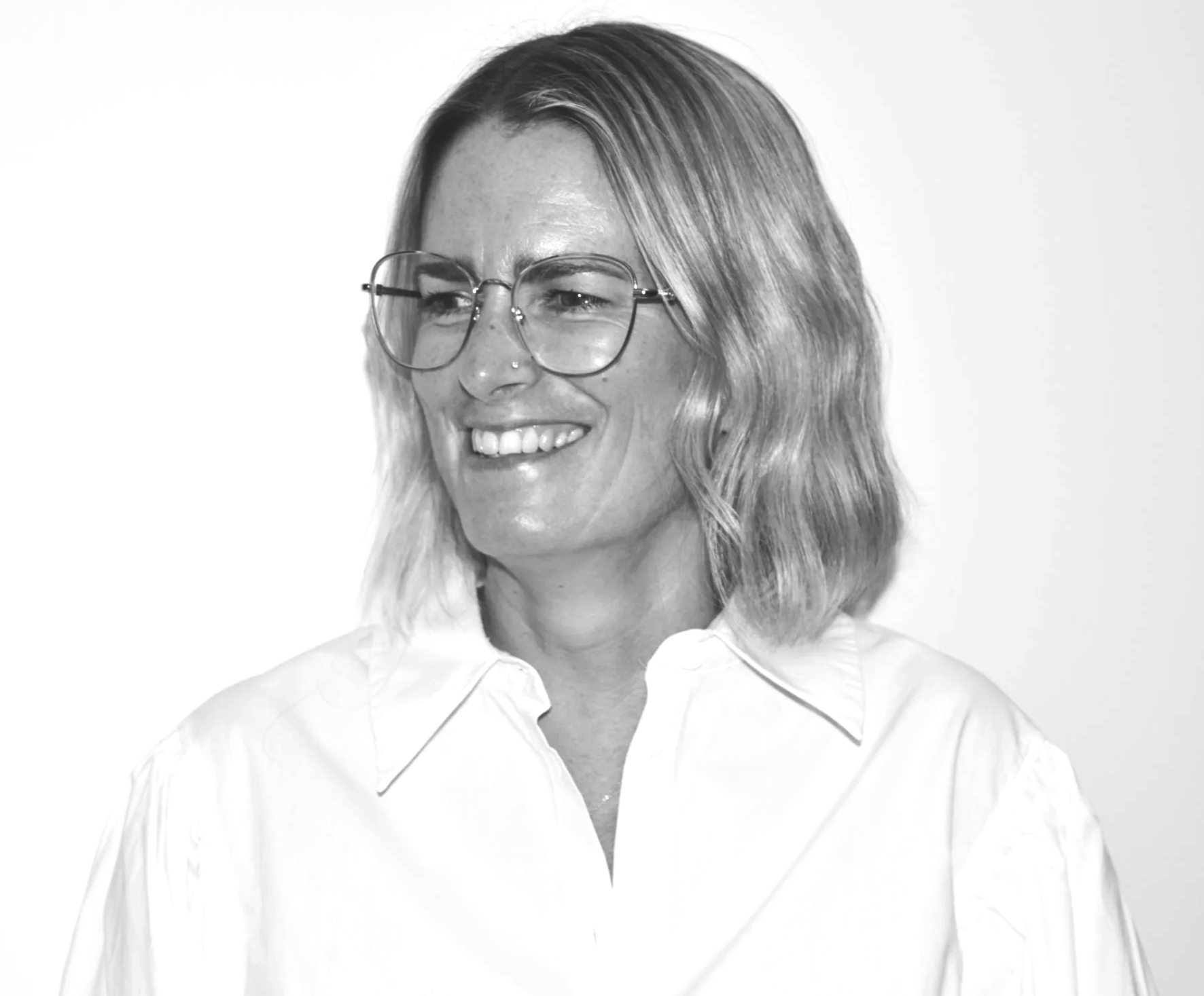Penny Piggott in a black and white portrait with shoulder-length hair, wearing glasses and a collared shirt, smiling slightly.