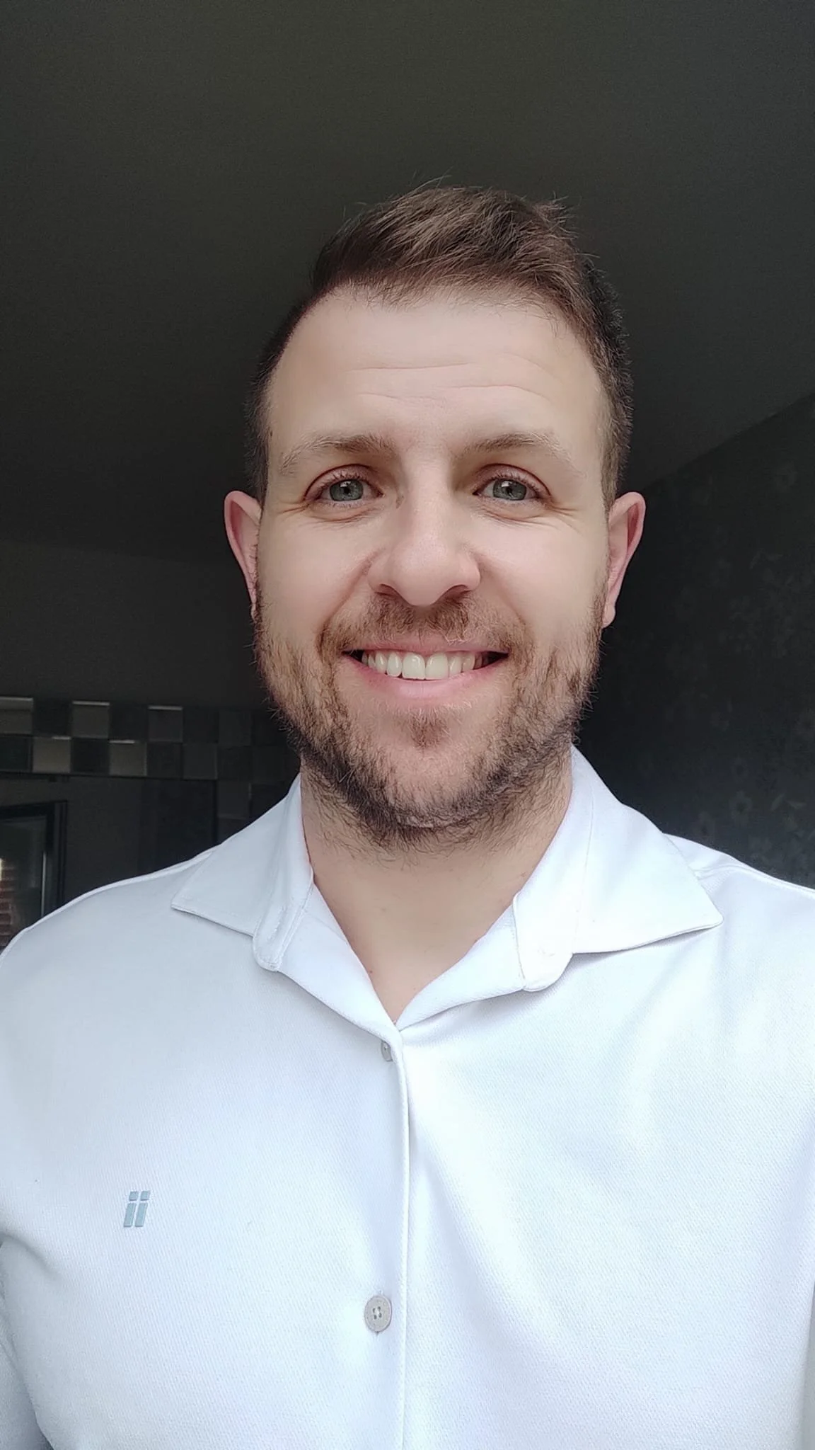 Retrato de un hombre joven con barba, cabello castaño, ojos azules y sonrisa, usando una camisa blanca, en un fondo oscuro.