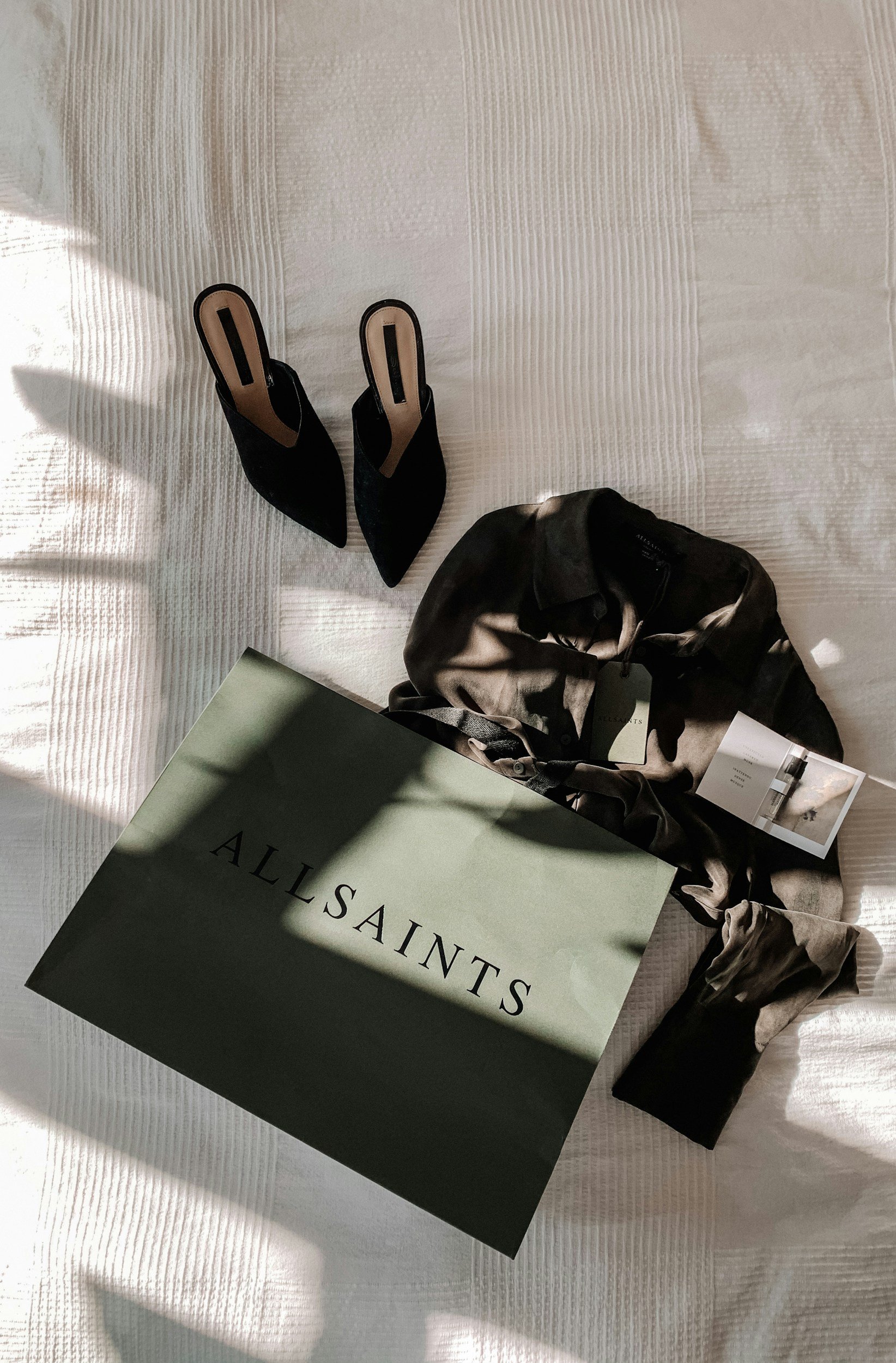 Zapatos negros de tacón alto, una bolsa de diseño y una caja de la marca Allsaints en una superficie blanca con sombra y luz natural.